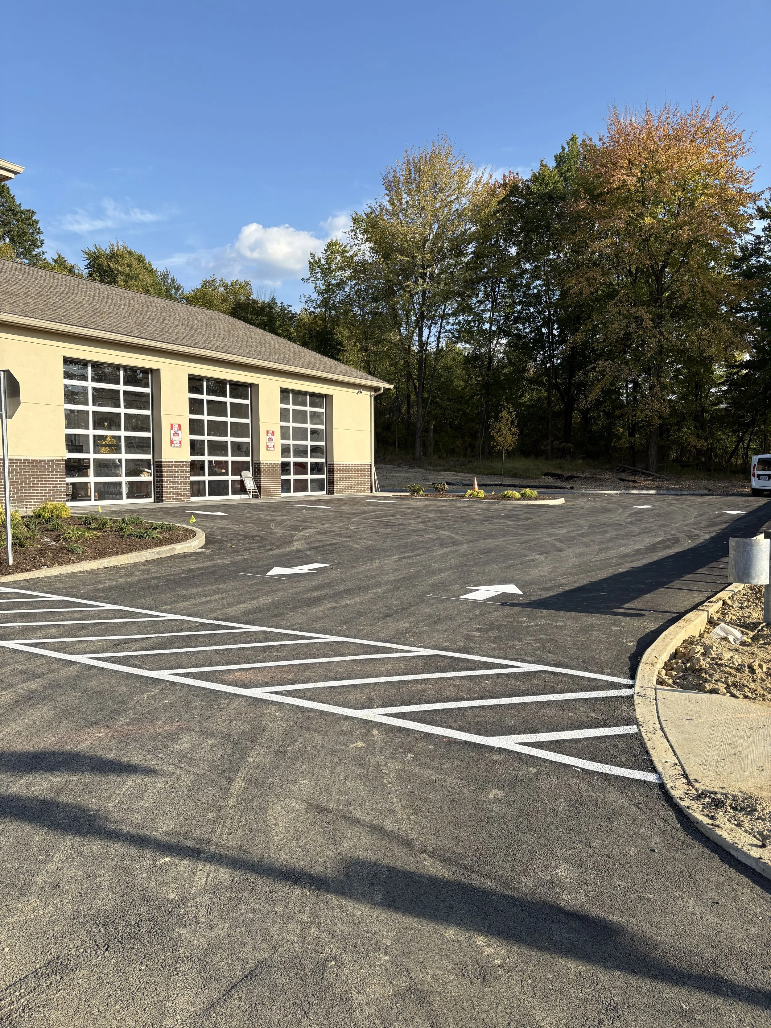 Empty parking lot with newly painted white lines and arrows, adjacent to a building with large garage doors, surrounded by trees with fall foliage, under a blue sky with some clouds.