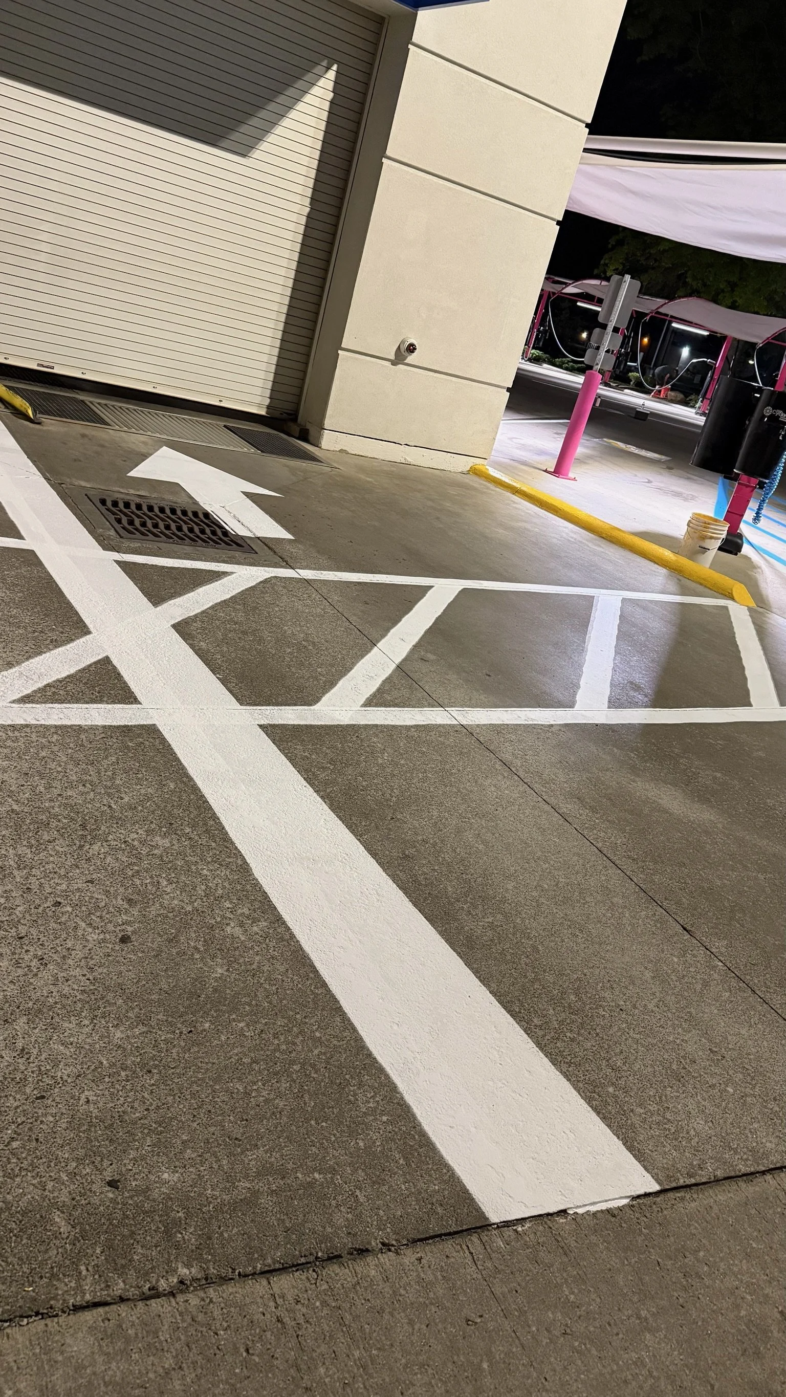 Empty parking space with white painted lines and a white arrow on the ground, next to a beige building with a closed roll-up door, and a yellow-painted curb. In the background, there are pink and black structures with tents or covers.