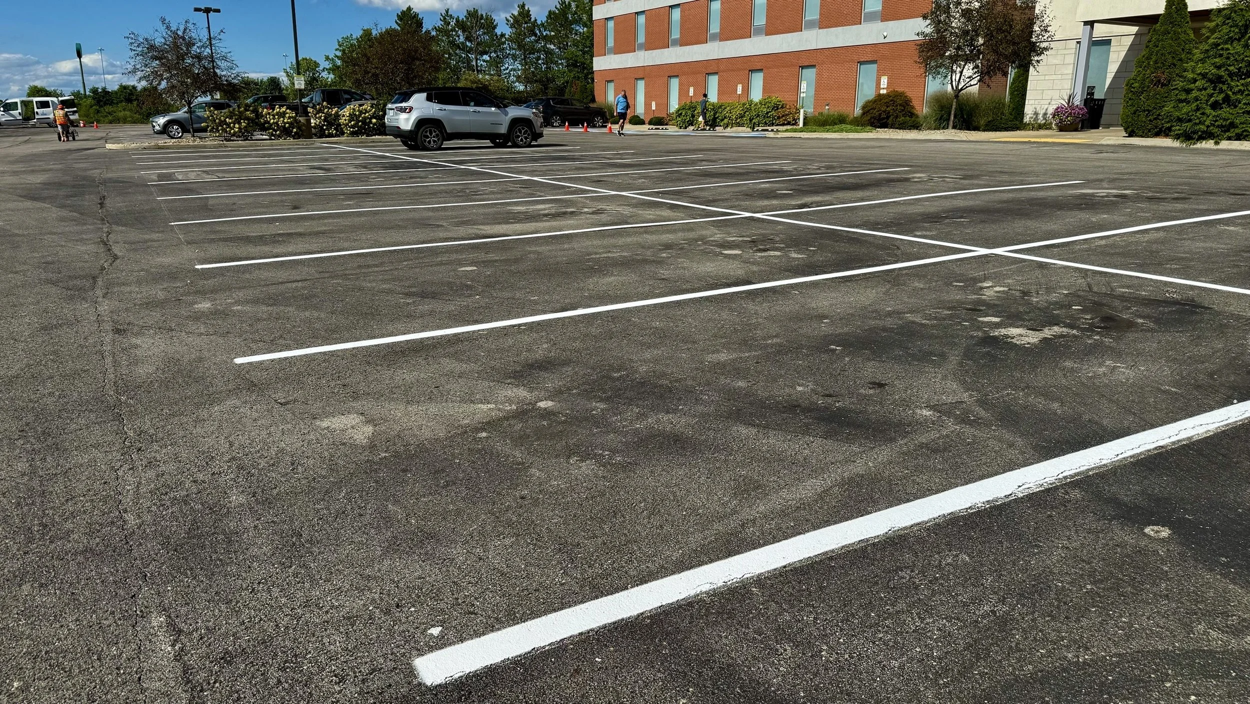Empty parking lot with a few cars parked near a building, with some people walking in the background.
