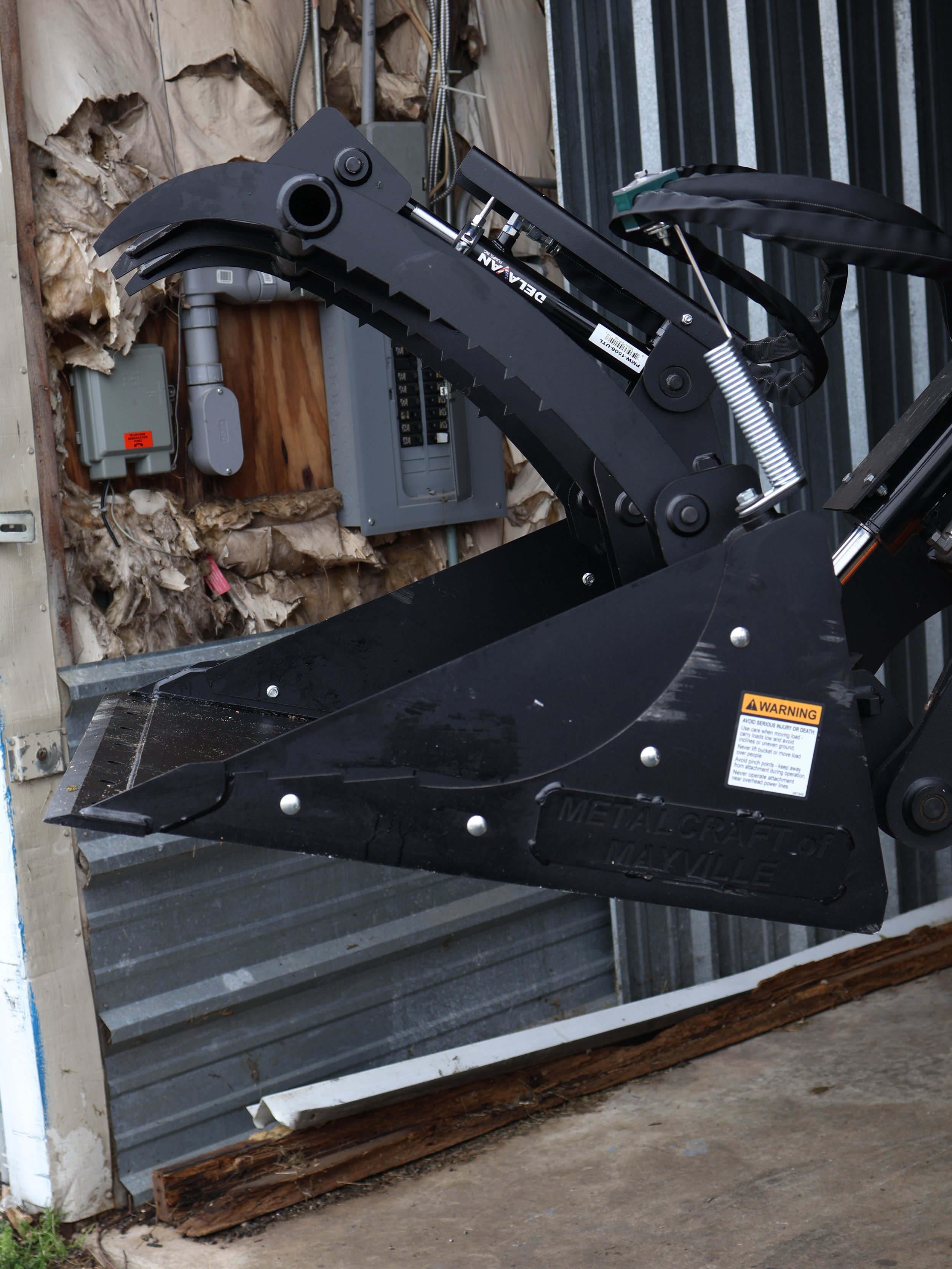 Close-up of a black Metalcraft snowplow attachment with hydraulic components in a garage.