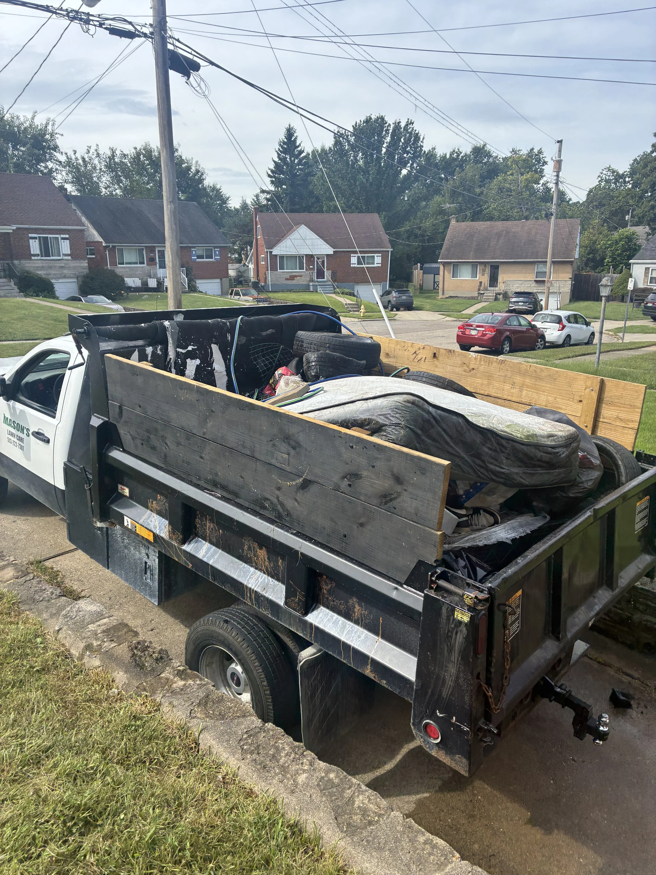 Landscaping truck with an open debris trailer parked on a residential street lined with brick houses, utility poles, and overhead wires.