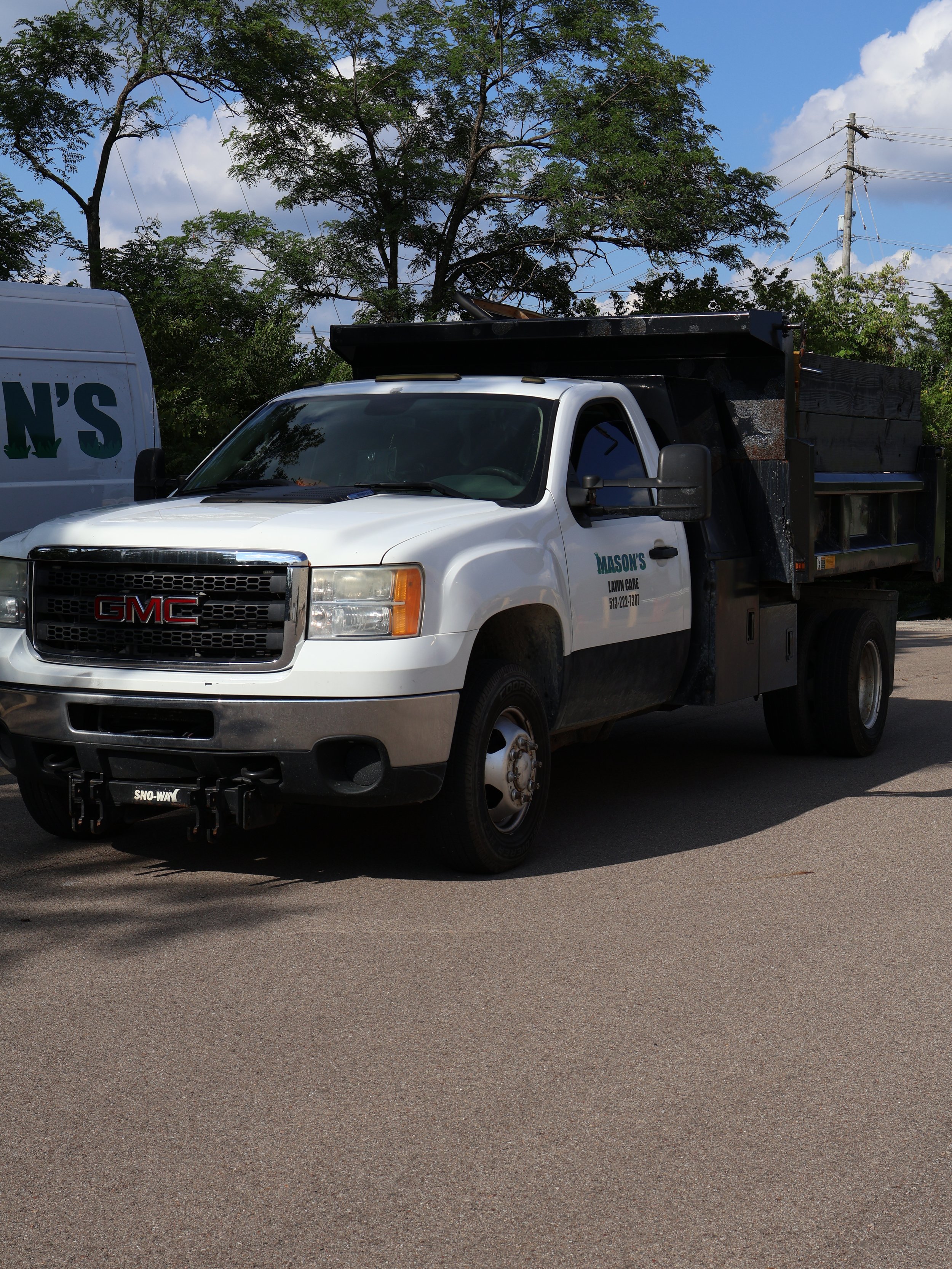 White GMC landscaping truck with a black dump bed parked beneath leafy trees and power lines.