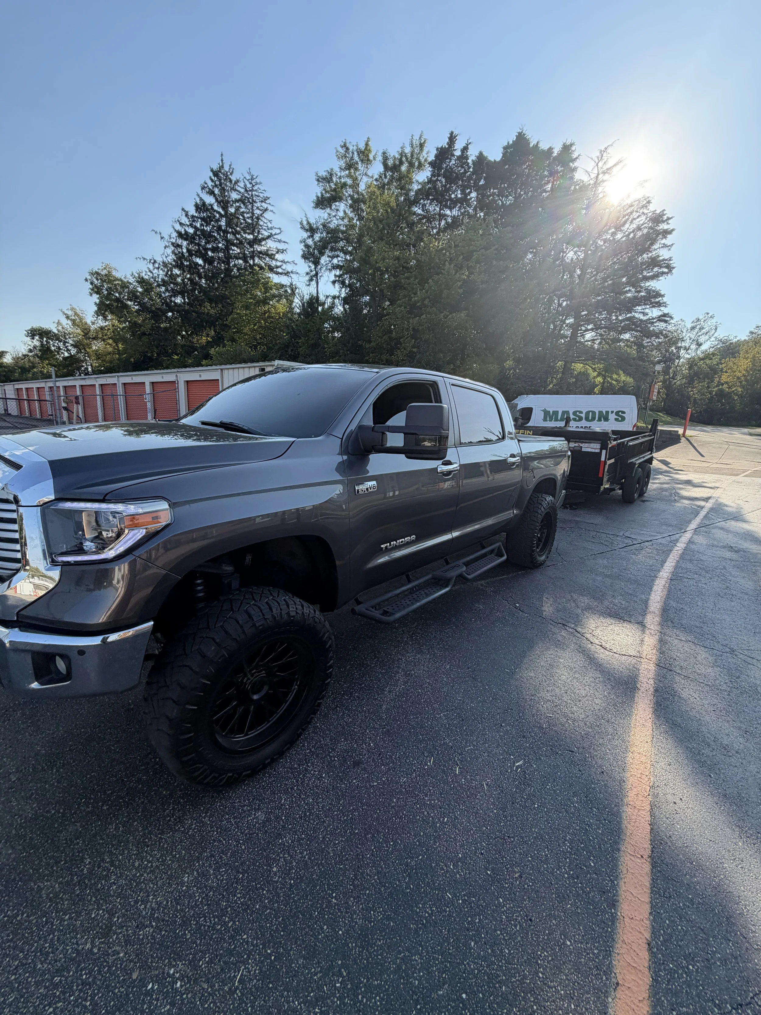 Dark gray Toyota Tundra pickup truck towing a utility trailer in a sunny parking lot, with storage units and trees in the background.