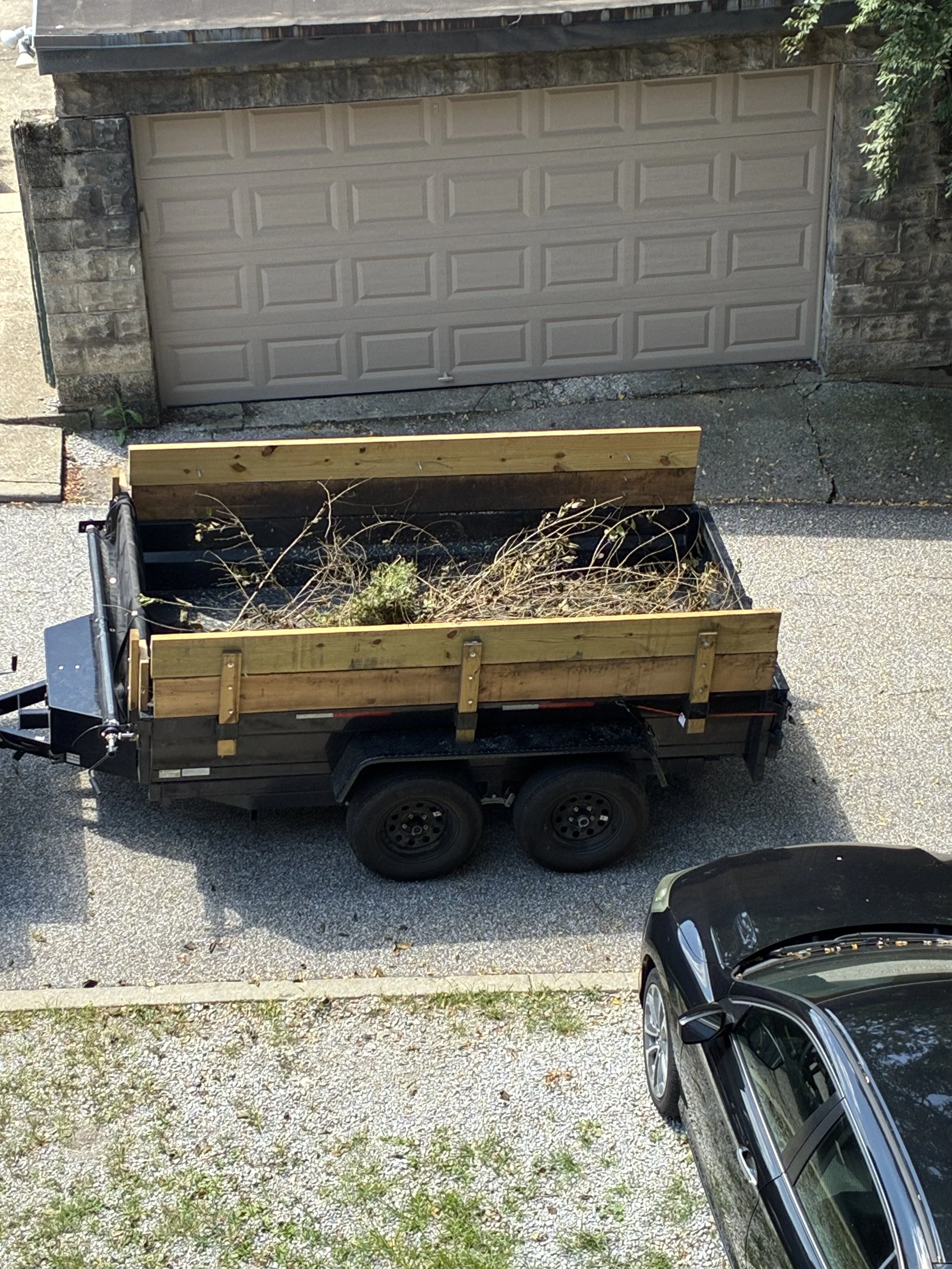 Utility trailer loaded with branches parked in front of a stone building and garage door.