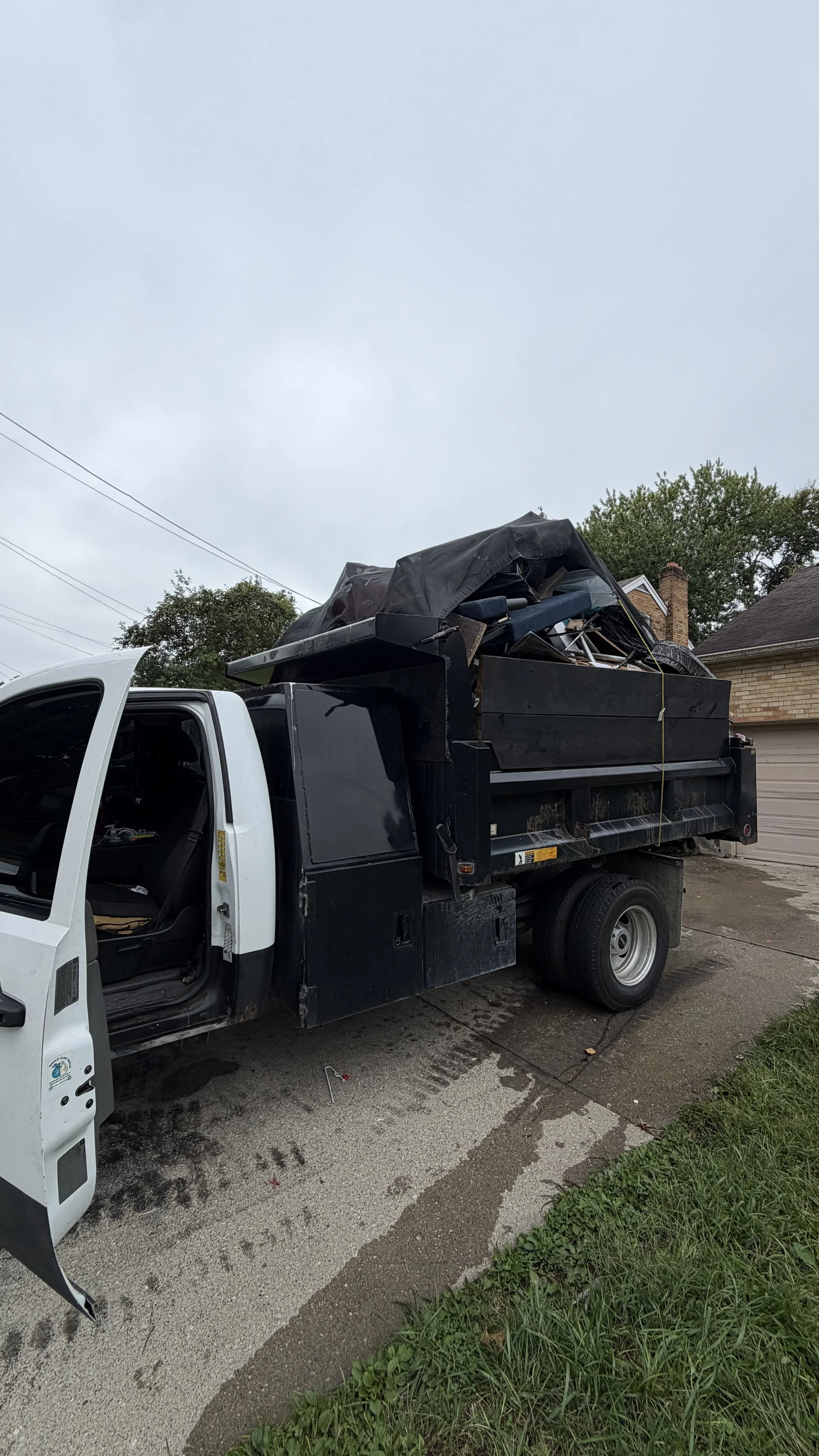 White box truck with an open door and black debris-filled dump bed parked in a residential driveway.