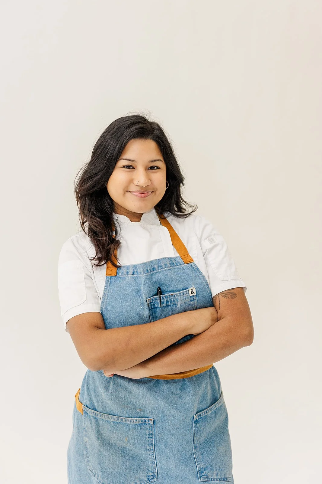 Young woman with shoulder-length dark hair wearing a gray t-shirt and denim apron with leather straps, standing in a modern kitchen.