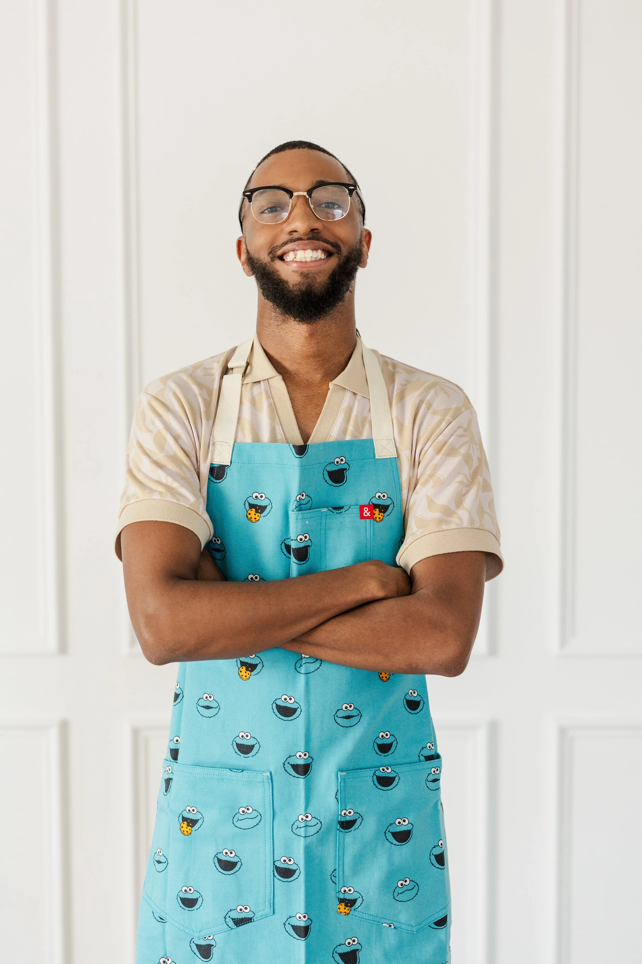 A man smiling with glasses and a beard, wearing a beige shirt and a blue apron with Sesame Street character faces, standing with arms crossed in front of a white wall.