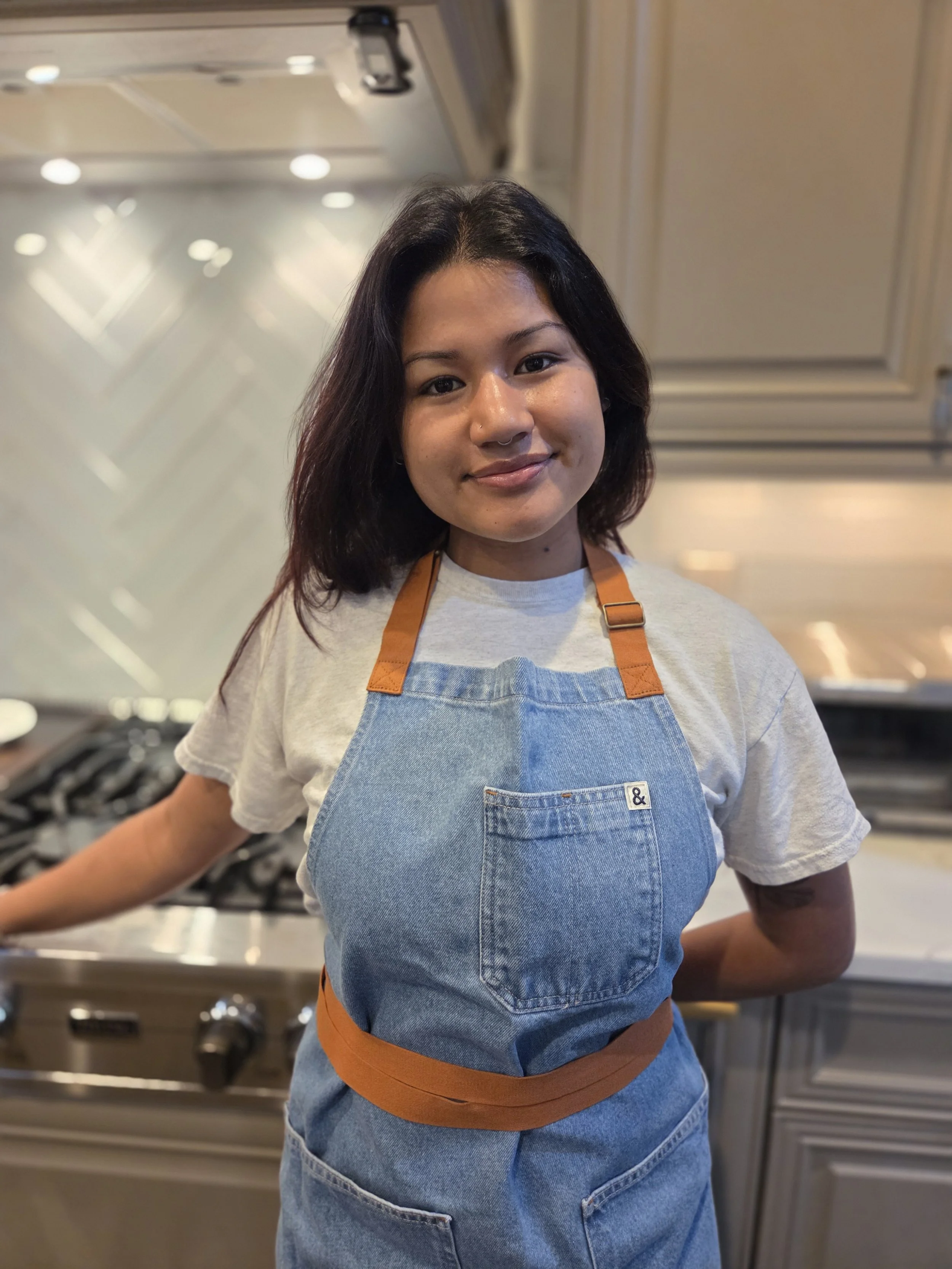 Young woman with shoulder-length dark hair wearing a denim apron over a gray t-shirt, standing in a modern kitchen.