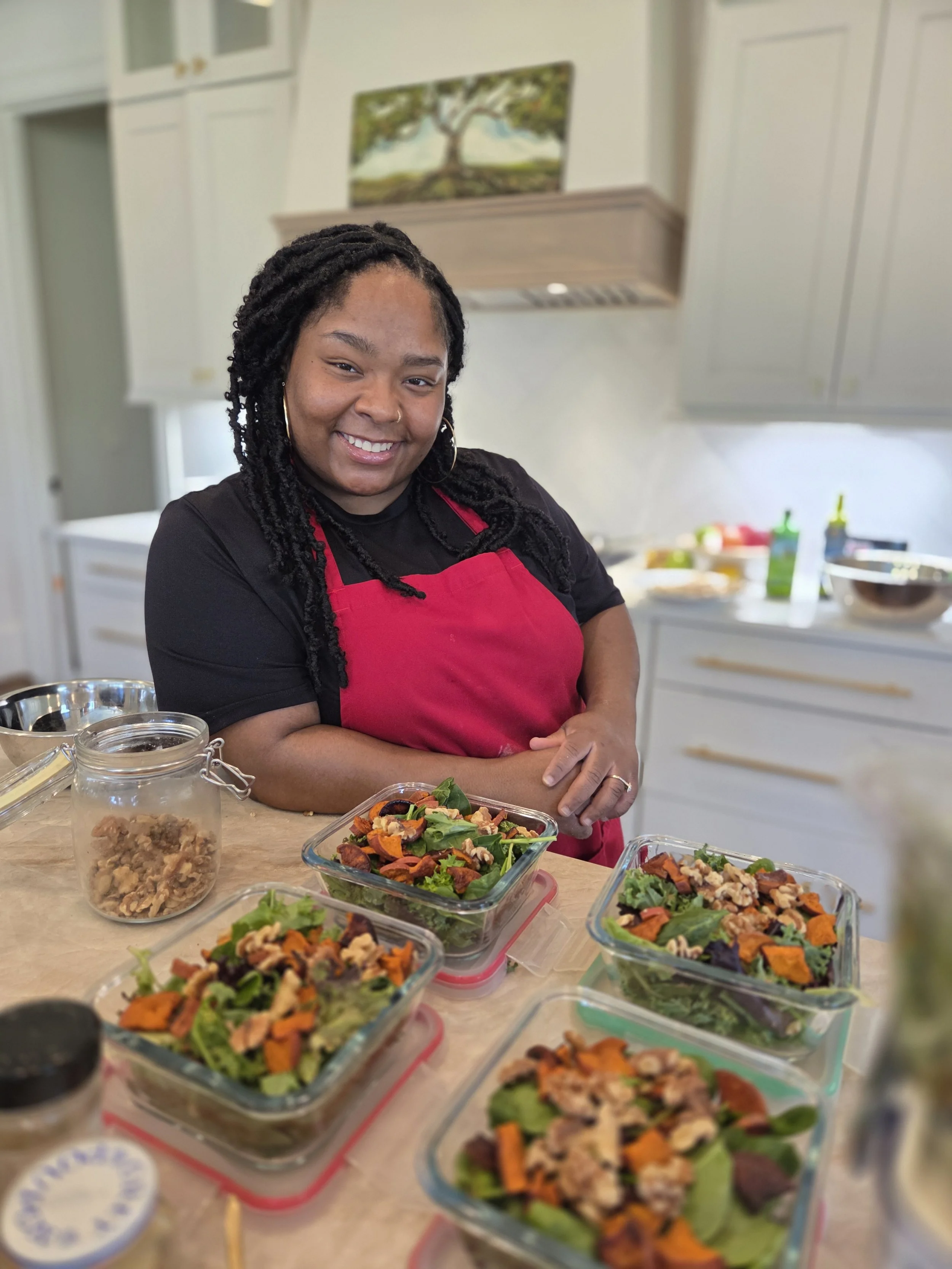 A woman wearing a black shirt and red apron stands in a kitchen with blond hair styled in twists, smiling at the camera, surrounded by several glass containers of salad with leafy greens, roasted carrots, walnuts, and other vegetables.