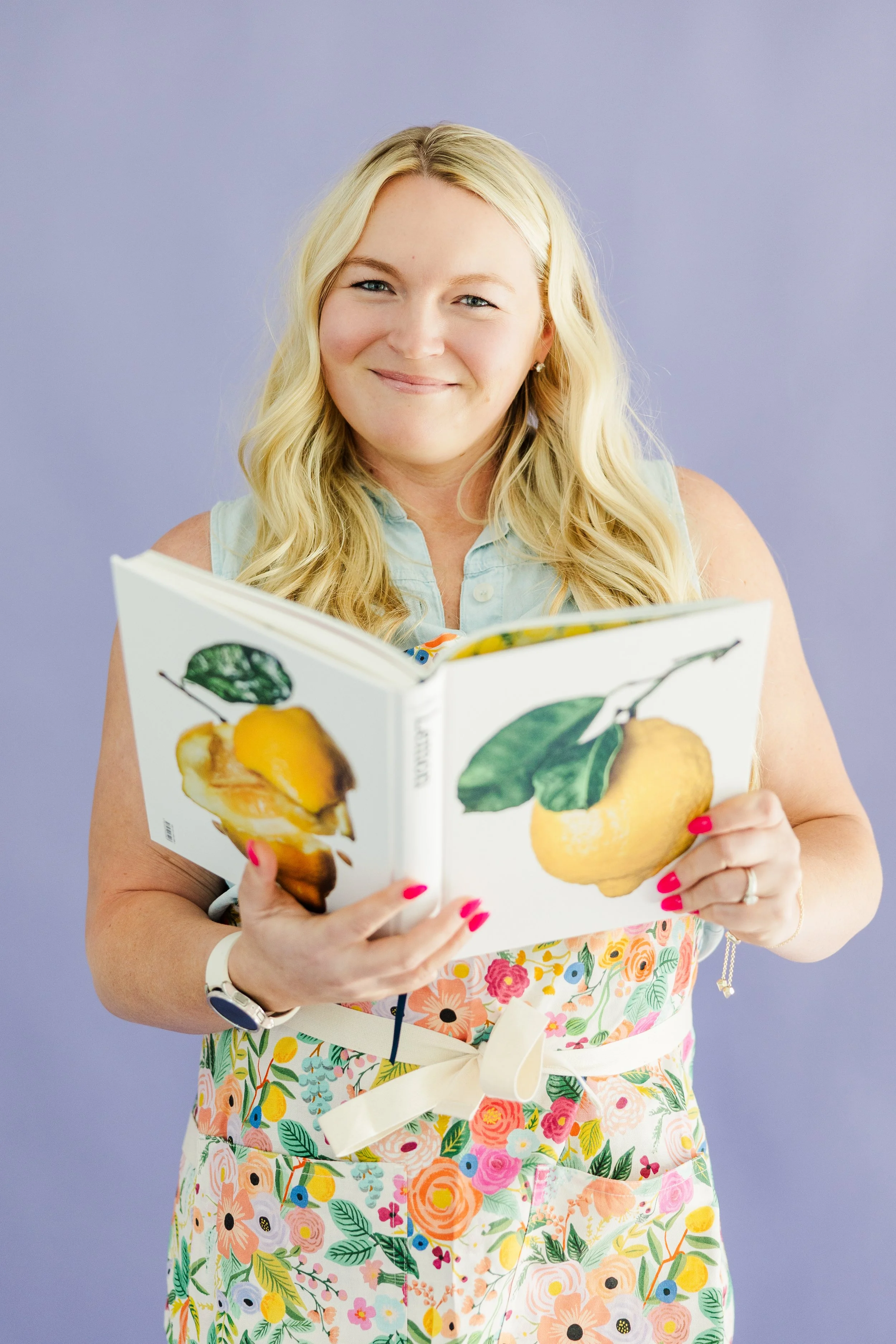 A woman with long blonde hair, smiling, holding an open cookbook with pictures of a lemon and a lemon with a leaf against a purple background.