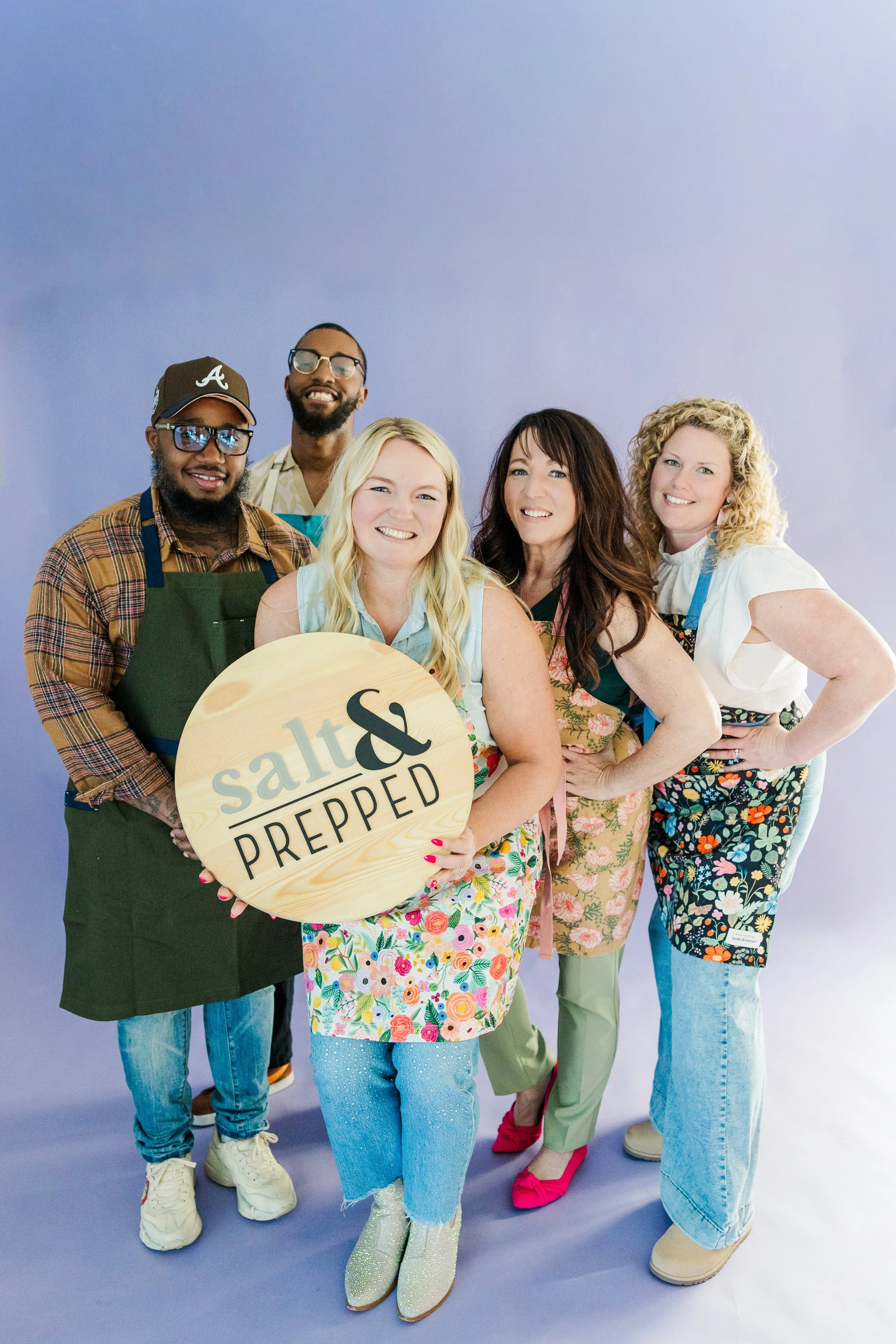Group of smiling diverse people wearing aprons, standing outdoors in front of a modern glass building.