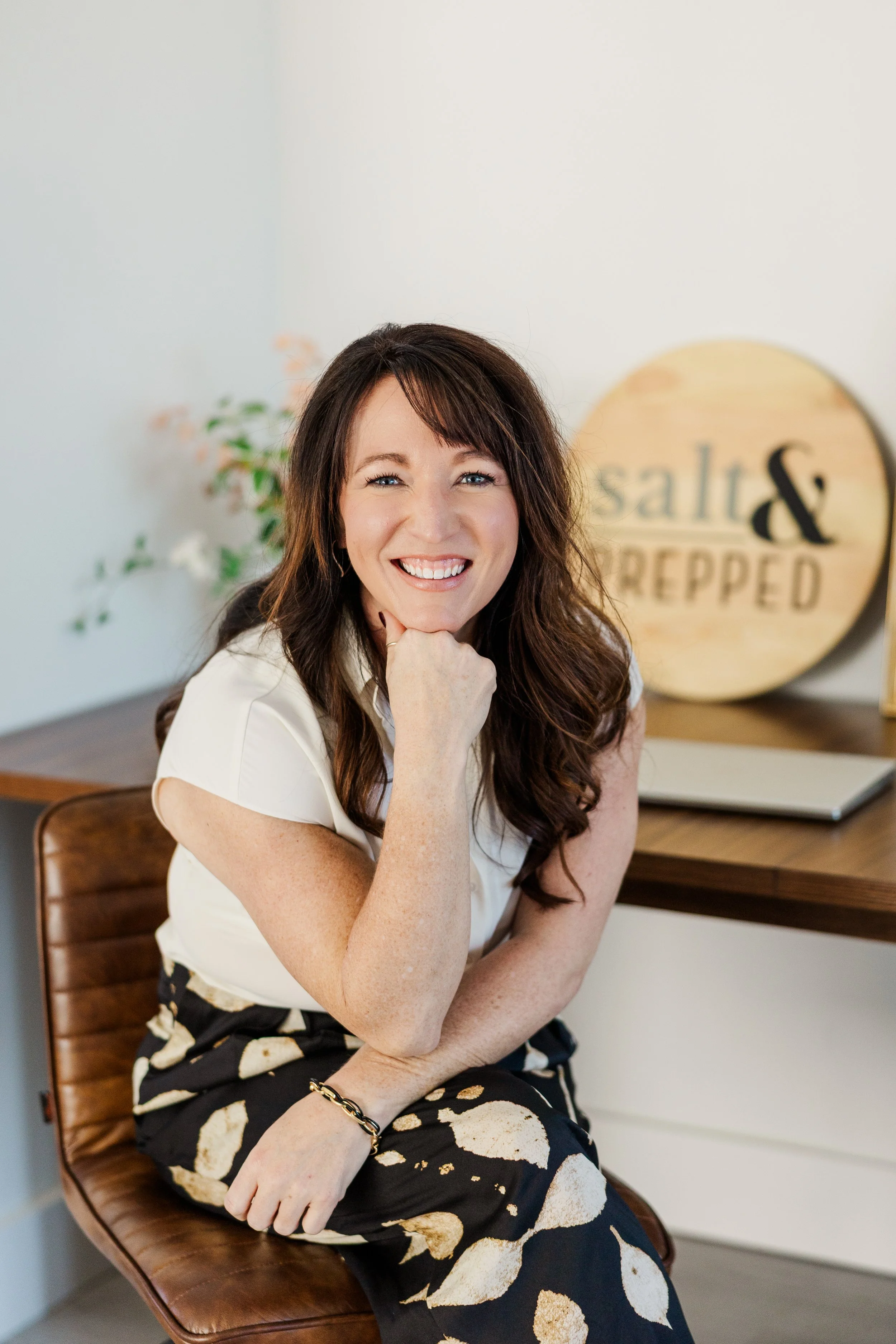 A woman with long brown hair sitting on a chair, smiling at the camera, with a sign that reads 'salt & rubbed' in the background.