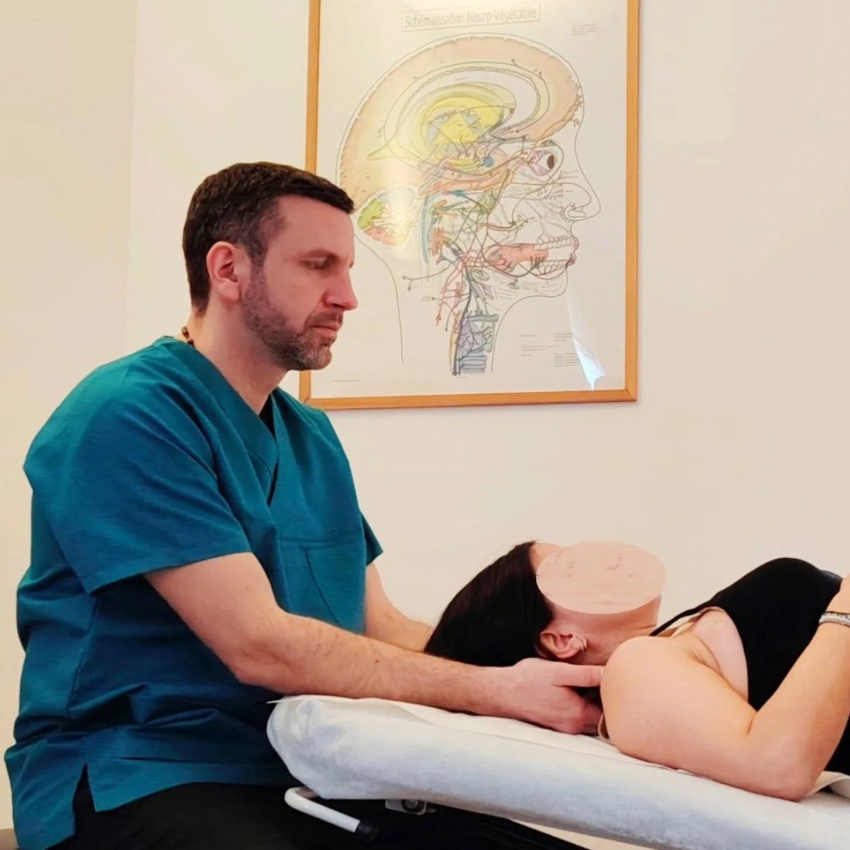 An osteopath adjusting a female patient lying face down on an examination table, in a osteopathy room with medical charts on the wall.