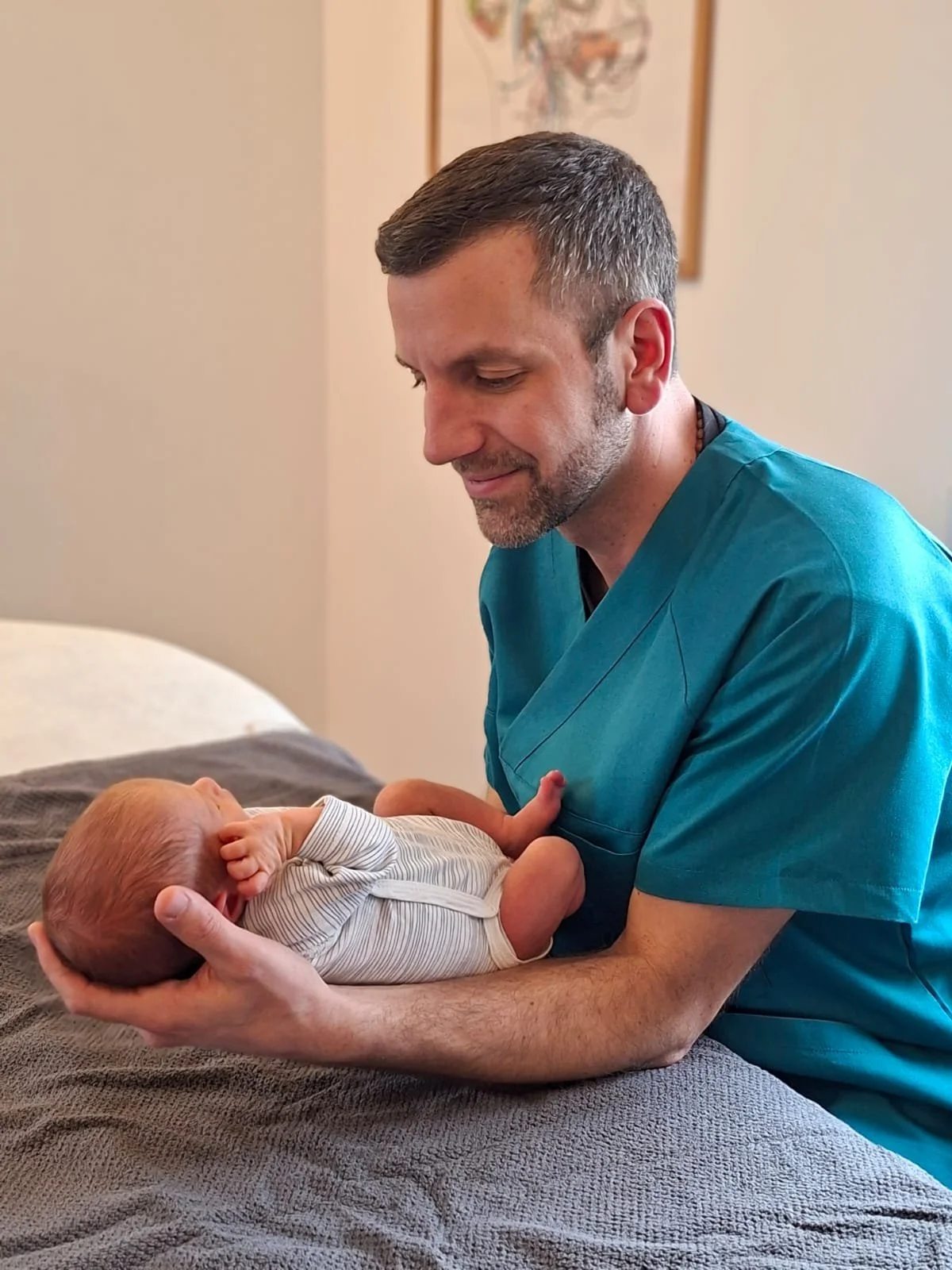 An Osteopath DO TO wearing teal medical scrubs holding a newborn baby in an osteopathy setting.