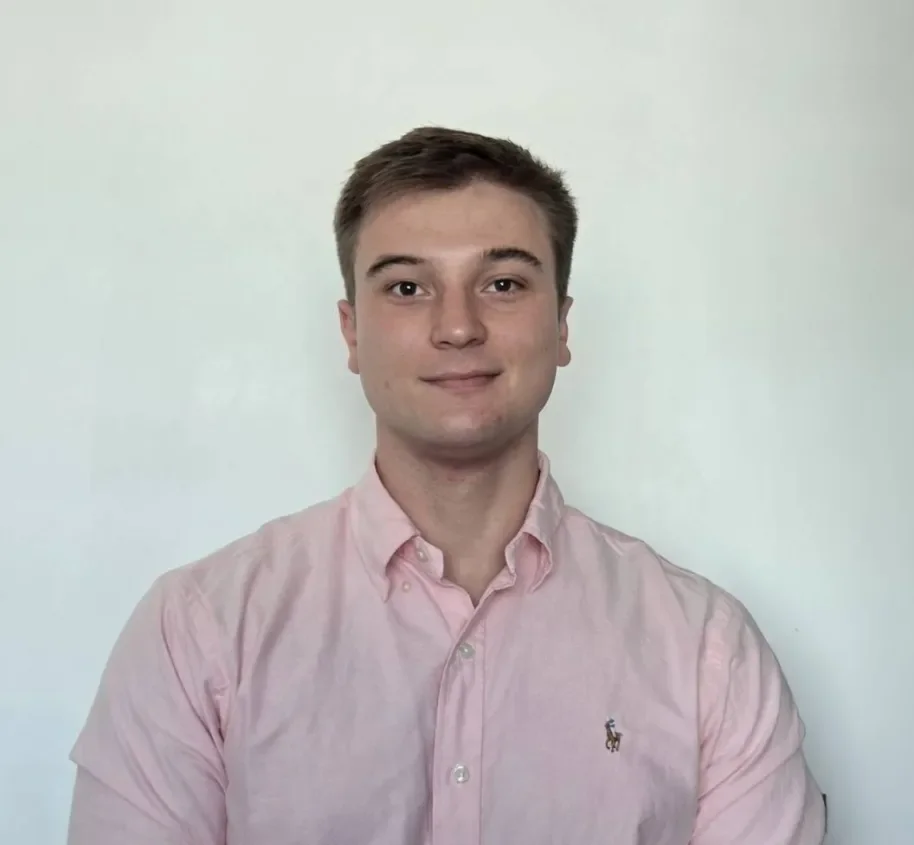 Young man with short brown hair wearing a light pink button-up shirt standing against a plain white wall.