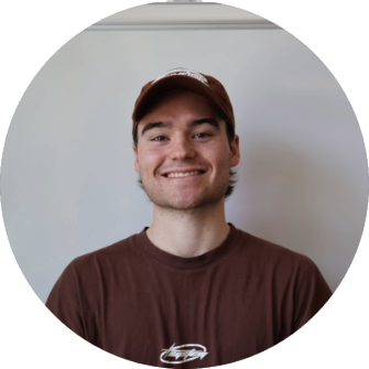 A young man with a brown cap and brown T-shirt smiling in front of a plain wall.