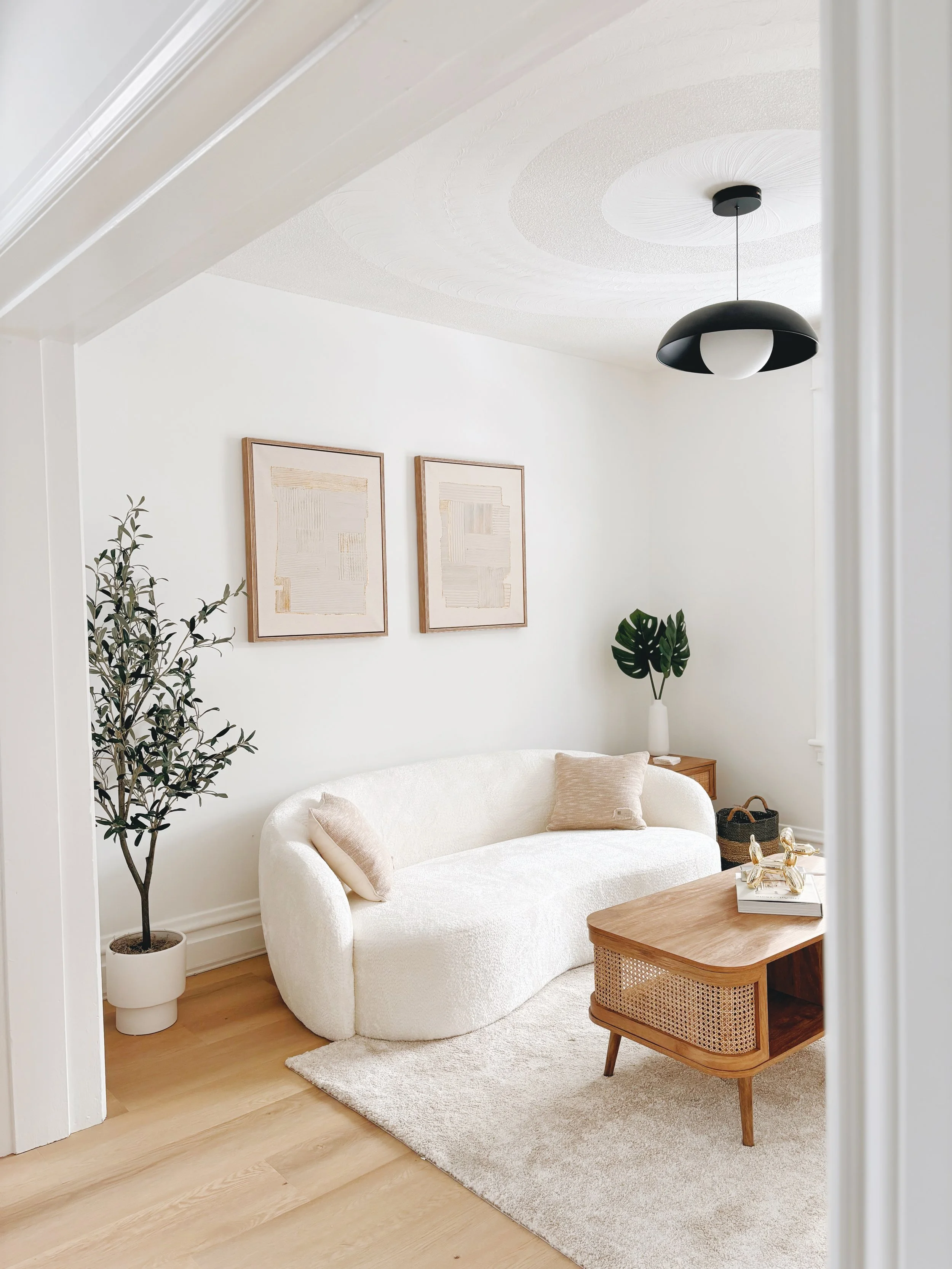 Minimalist living room with a white curved sofa, wooden coffee table, indoor plant, geometric wall art, and a modern light fixture.