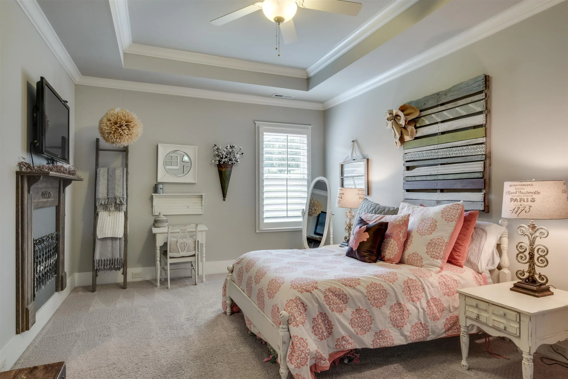 Cozy bedroom with pink floral bedding, rustic wood paneling above the bed, decorative pillows, bedside table with ornate lamp, wall-mounted TV, ceiling fan, and window with shutters. Kevin Martelli Broker and Realtor at Advise Nashville Onward Real 