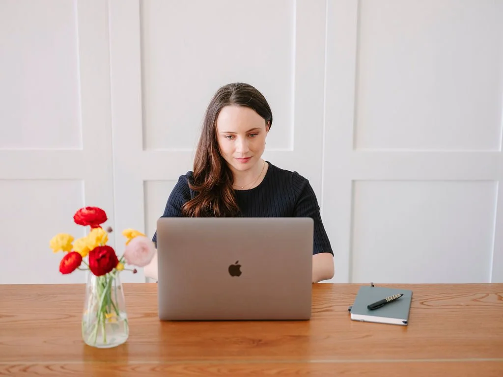 A young woman with long brown hair working on a laptop at a wooden desk with a vase of flowers, a notebook, and a pen.