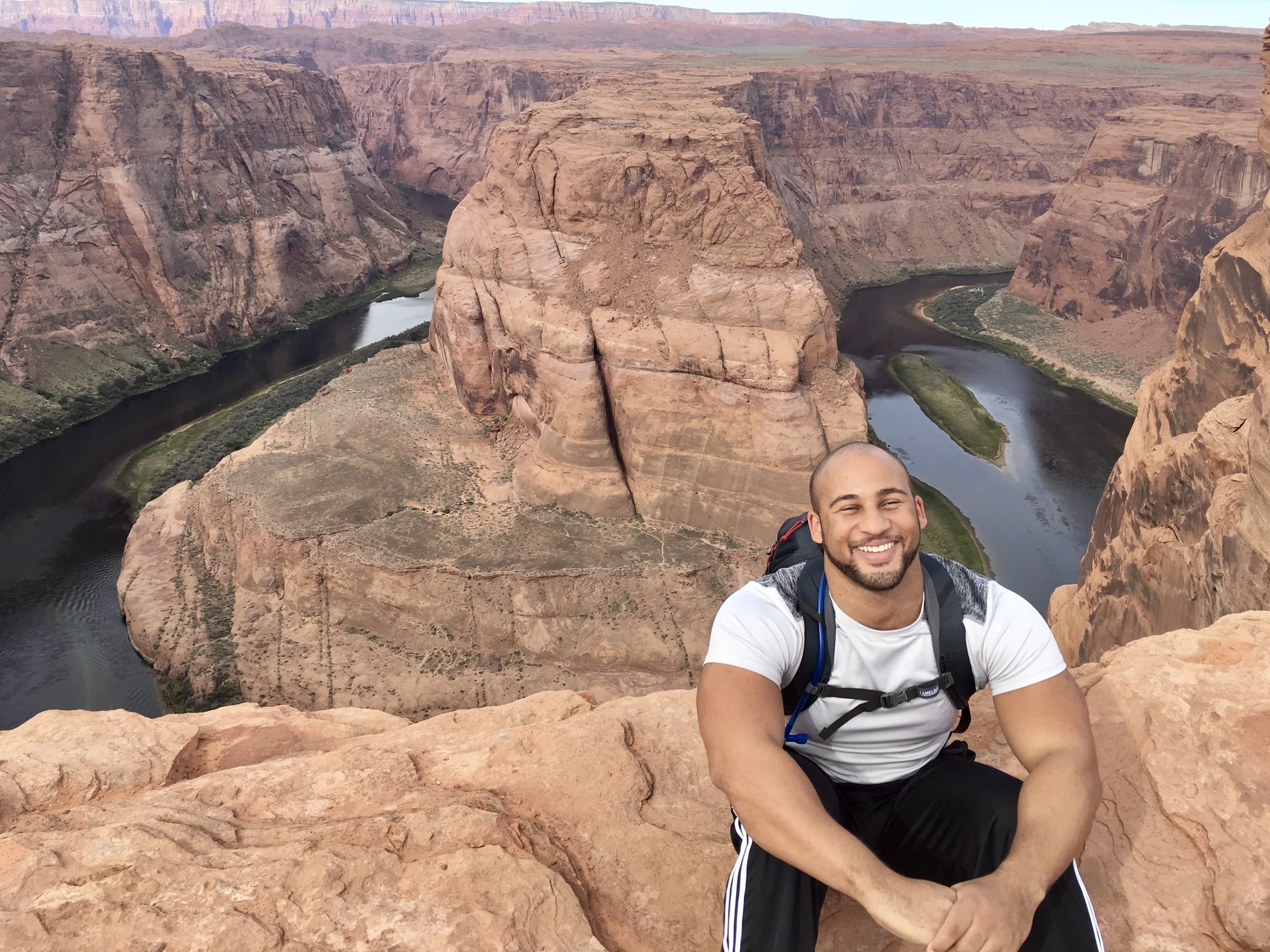 A man smiling and sitting on a rocky ledge overlooking the Colorado River at Horseshoe Bend in Arizona.