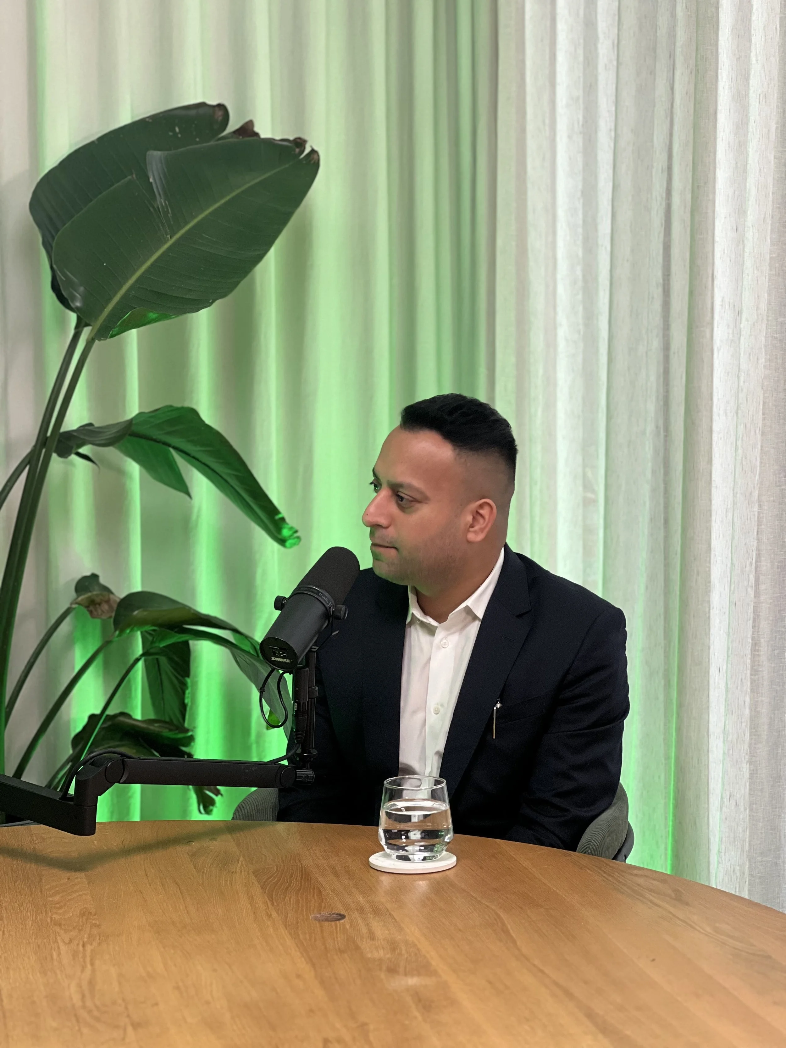 A man in a suit sitting at a wooden table with a glass of water, a microphone, a lush green plant, and a white curtain backdrop.