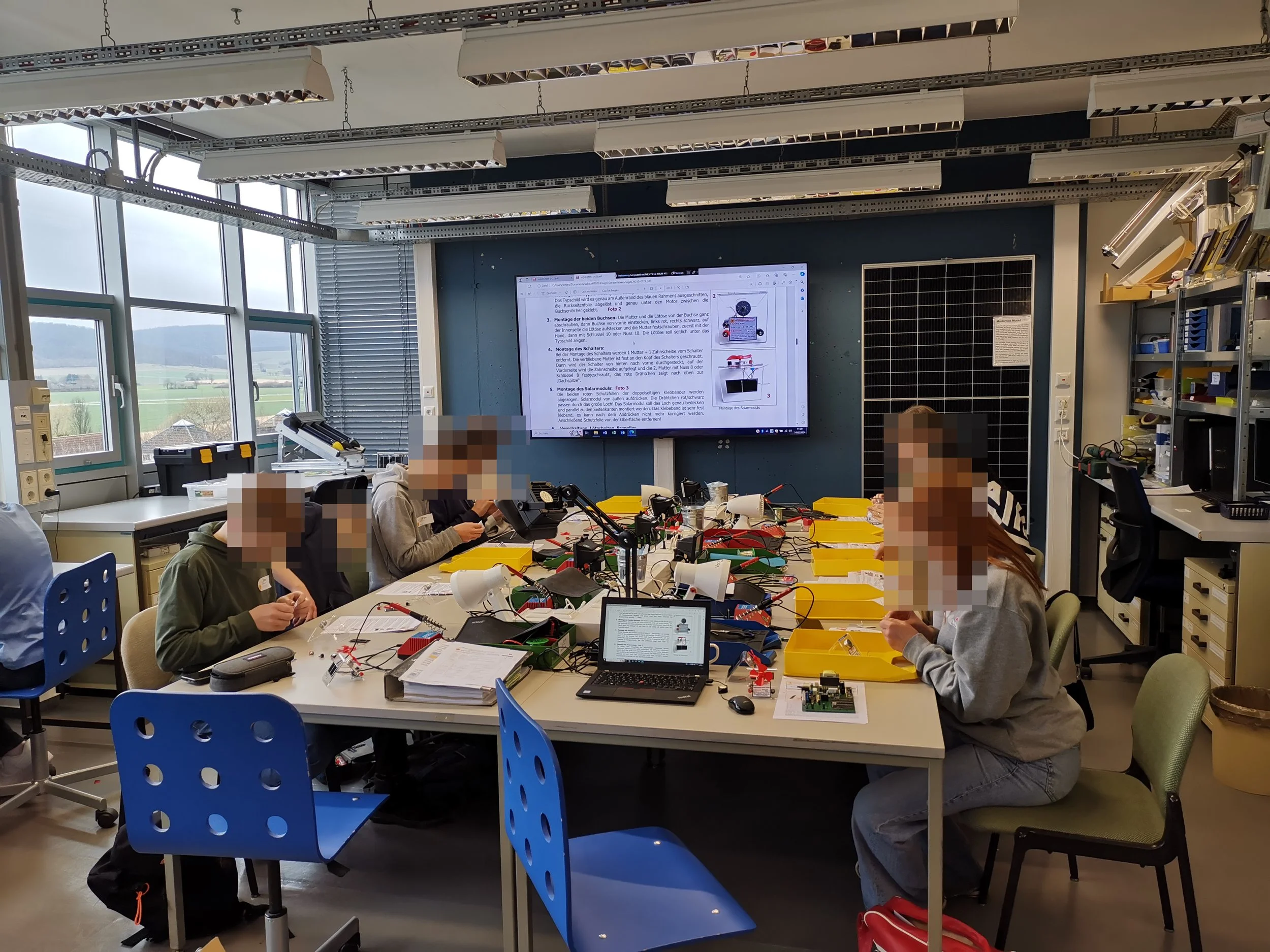 Ein Gruppenfoto von mehreren Schülern in einem Technikraum. Sie arbeiten an elektronischen Experimenten mit Schaltkreisen und Werkzeugen, umgeben von Computern, Arbeitsmaterialien und einer großen digitalen Anzeige an der Wand.