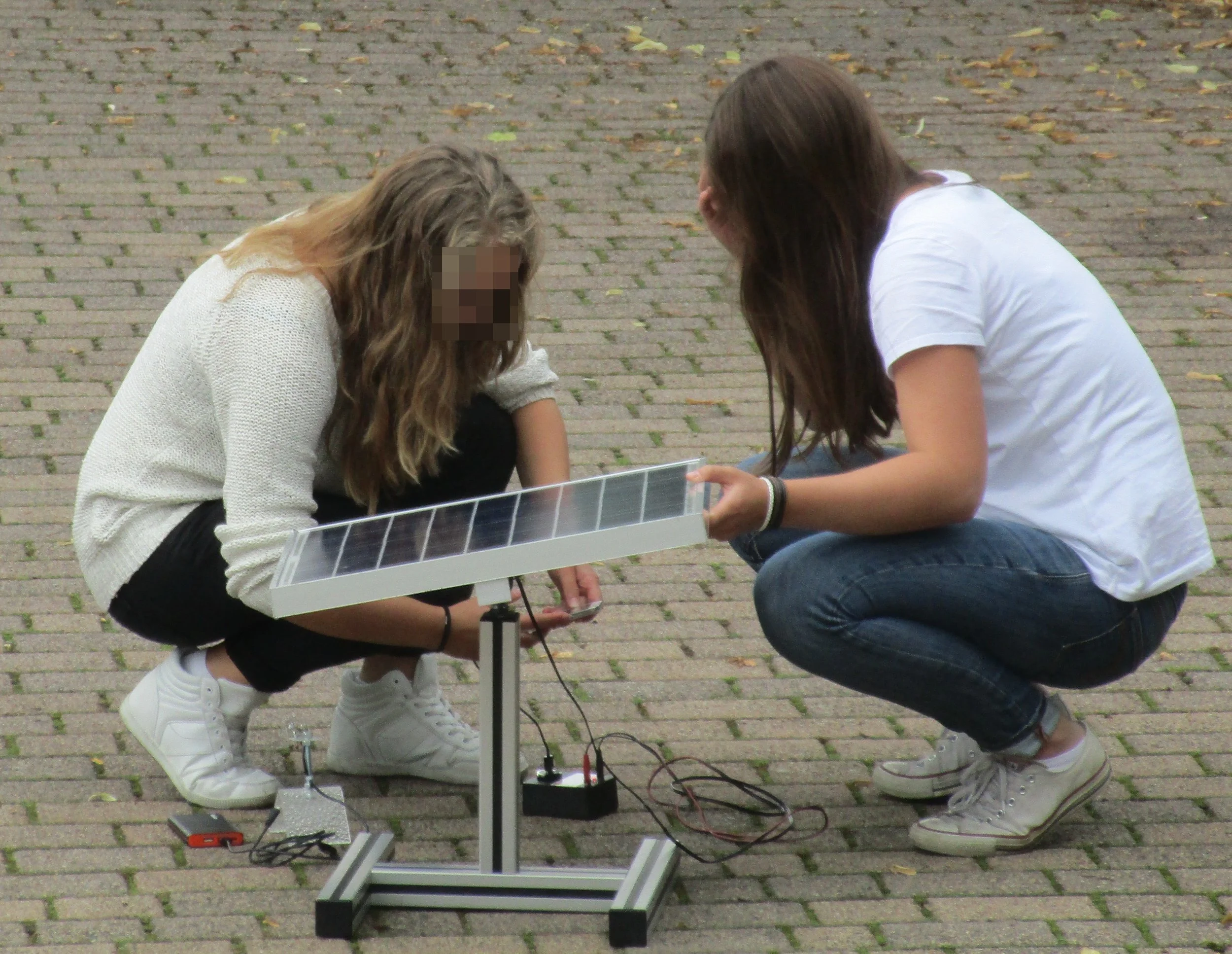 Zwei junge Frauen arbeiten im Freien an einem wissenschaftlichen Experiment mit einer solarbetriebenen Vorrichtung auf einem gepflasterten Boden.