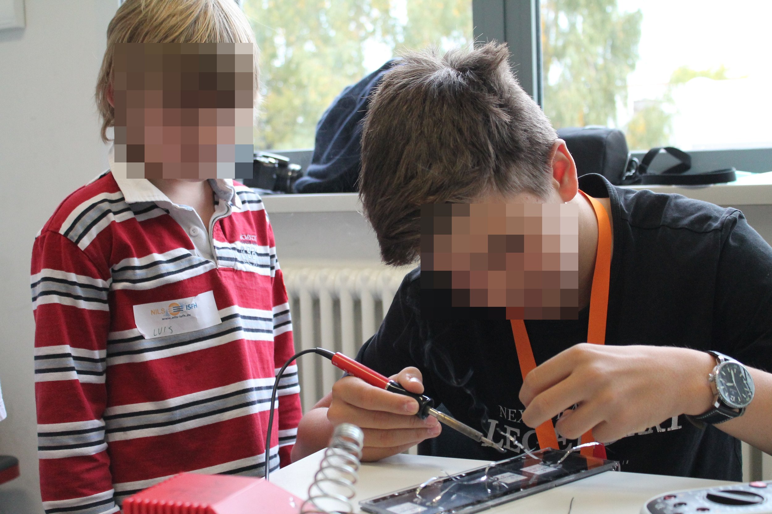 Ein Junge arbeitet mit einem Lötkolben an einem elektronischen Projekt, während ein anderer daneben zuschaut, in einem Klassenraum mit großen Fenstern im Hintergrund.