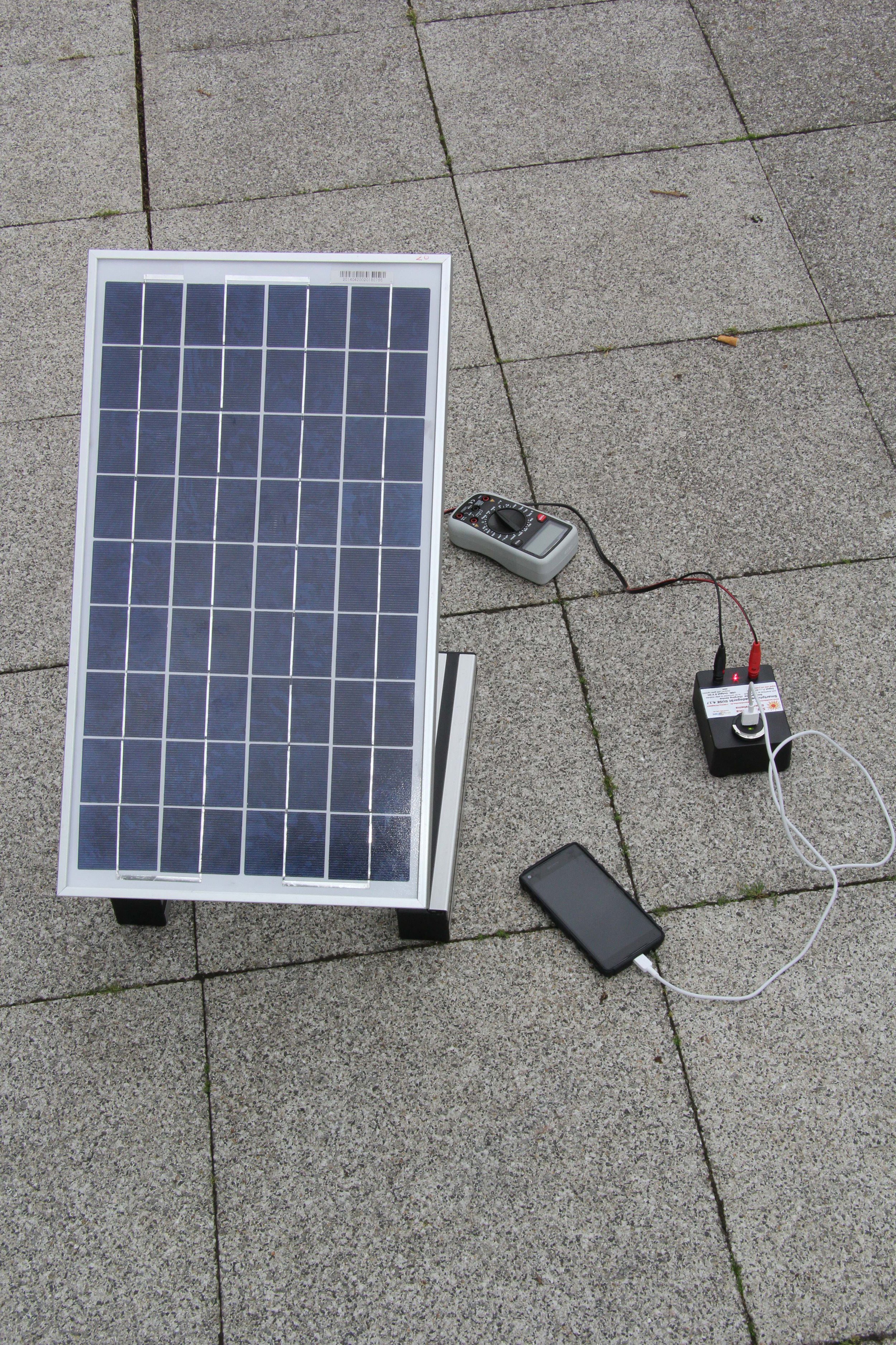 Solarmodul, verbunden mit einem Multimeter, einem Akku und einem Smartphone, auf einer Gehwegplatte im Freien.
