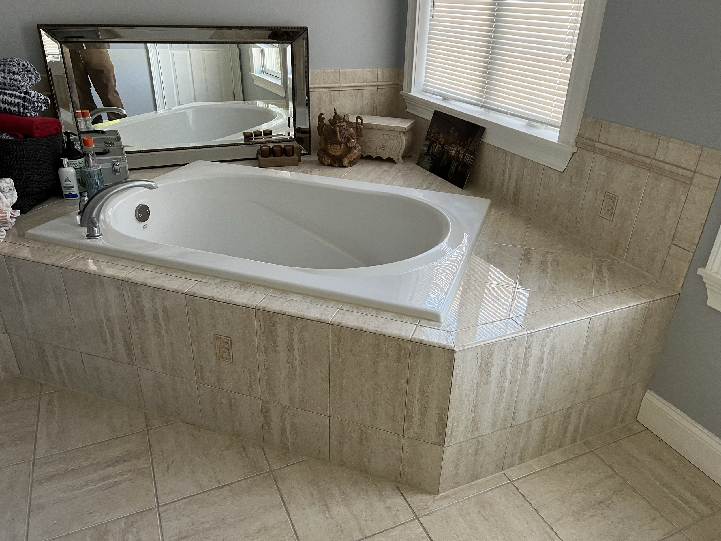 A bathroom with a large white bathtub surrounded by beige tiled countertops and walls, a window with blinds, and a mirror reflecting part of the room.