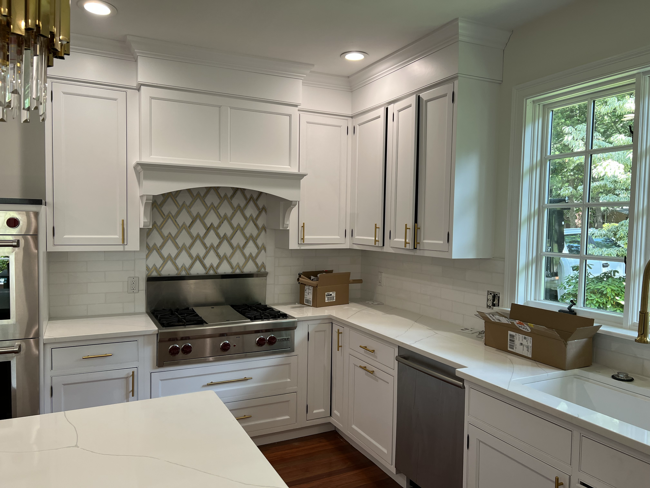 Modern kitchen with white cabinets, gold handles, marble countertops, a stainless steel stove, and a large window showing green foliage outside.