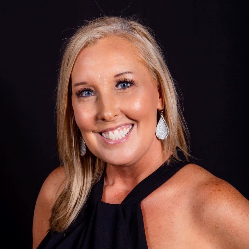 A woman with blonde hair and blue eyes smiling, dressed in a black top with silver earrings, against a black background.