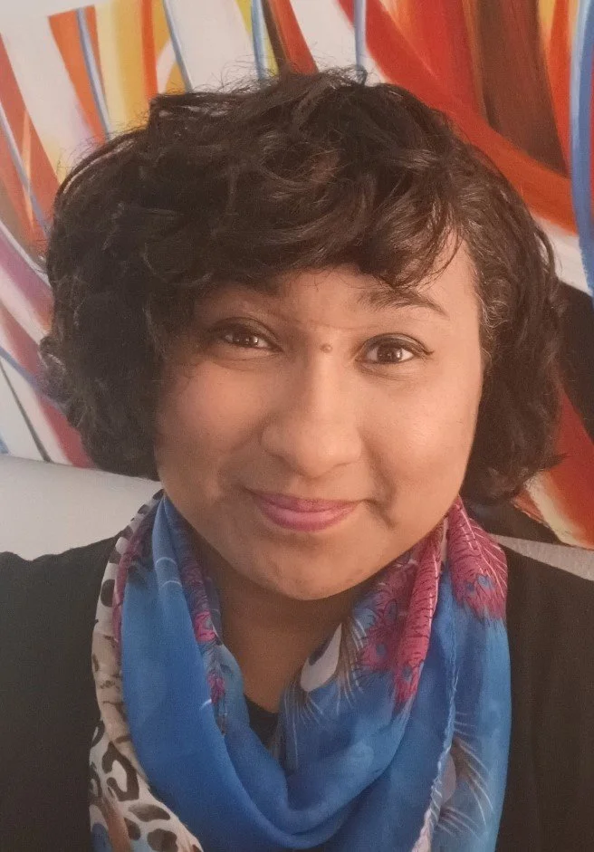 A woman with short, curly dark hair smiling in front of a colorful, abstract background, wearing a blue scarf with reddish-pink accents.