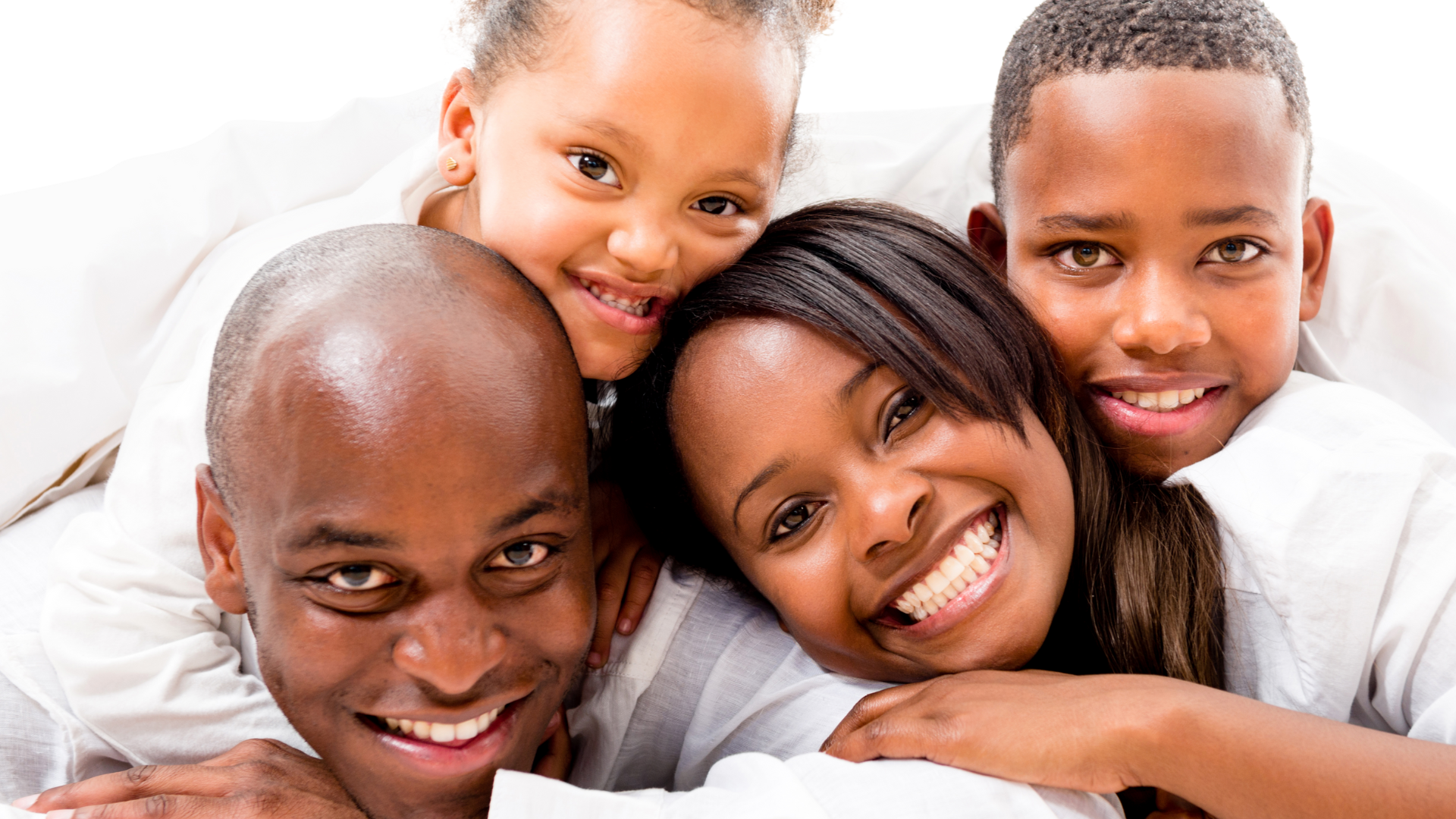 A happy multi-ethnic family of four, including two adults and two children, lying on a bed, smiling and closely cuddling together.