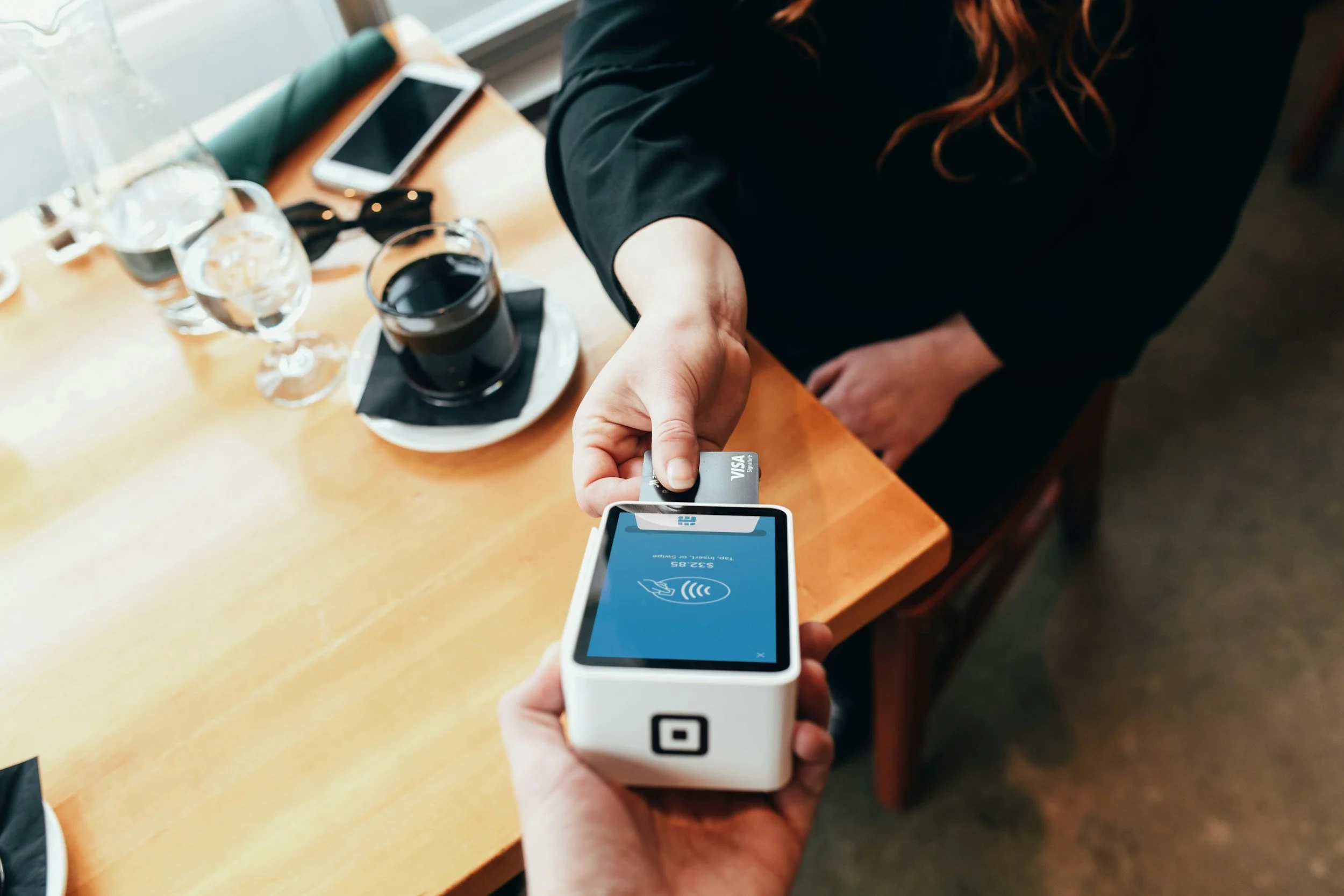 Person using a contactless payment terminal to pay with a card at a coffee shop. There are glasses of water, a glass of dark beverage, a smartphone, and other items on the table.