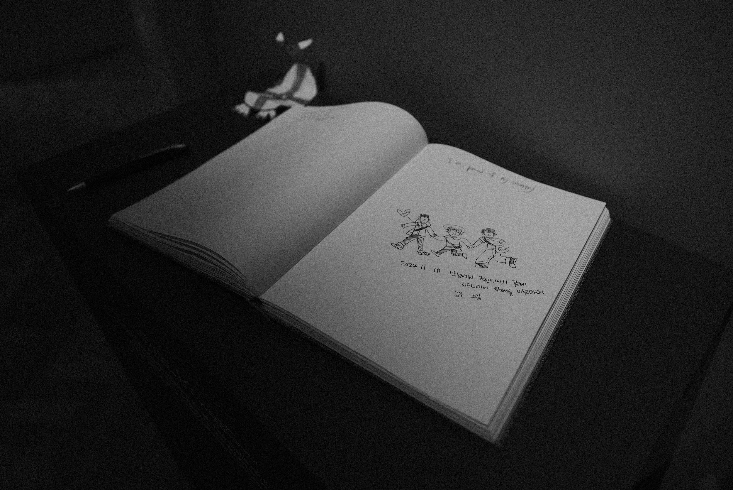 Open sketchbook with a drawing of three people holding hands, with a heart and a star; handwritten notes and date on the page, in a dimly lit setting.