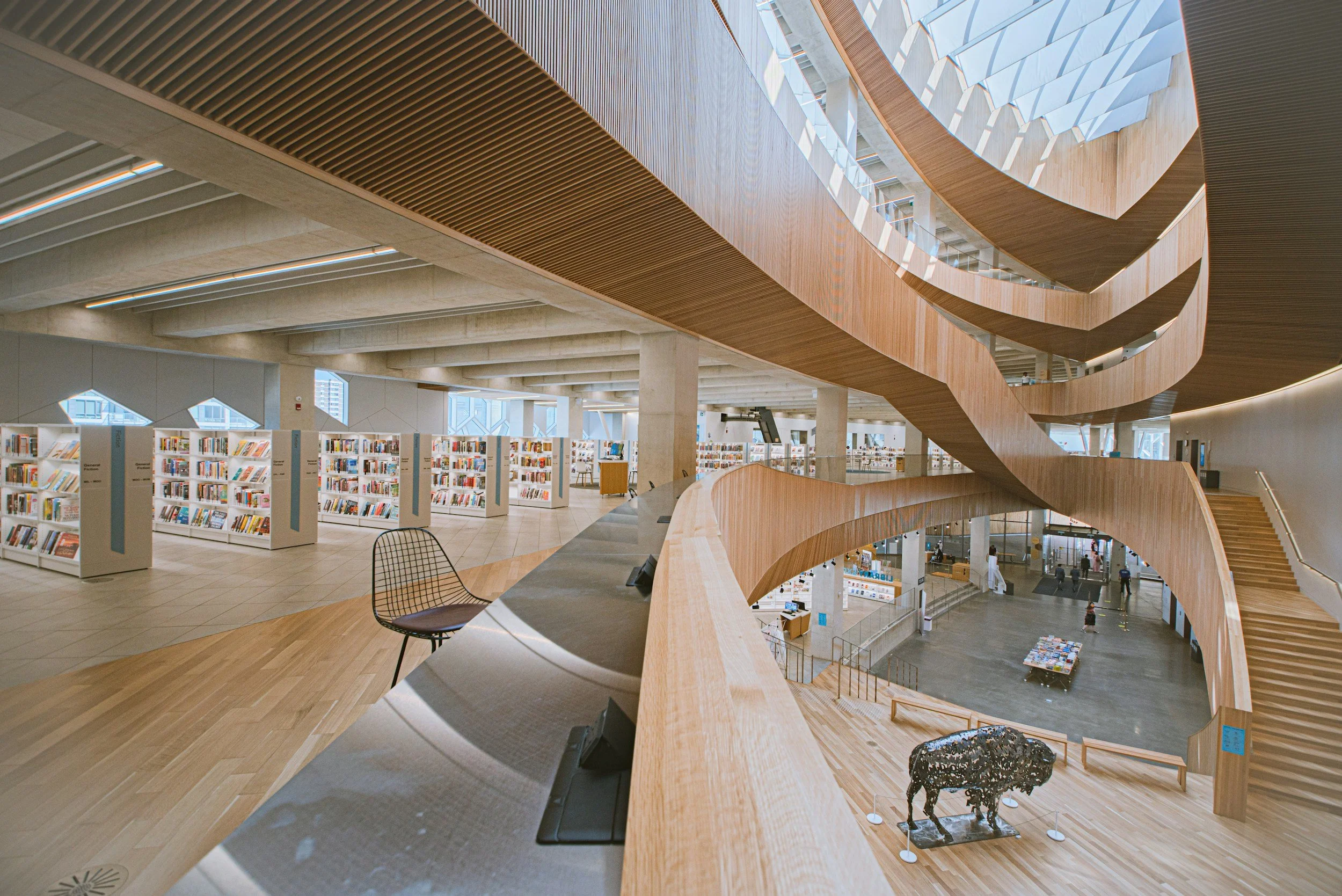 Interior of a modern library with bookshelves, a curved wooden staircase, and seating areas illuminated by natural light from large skylights.