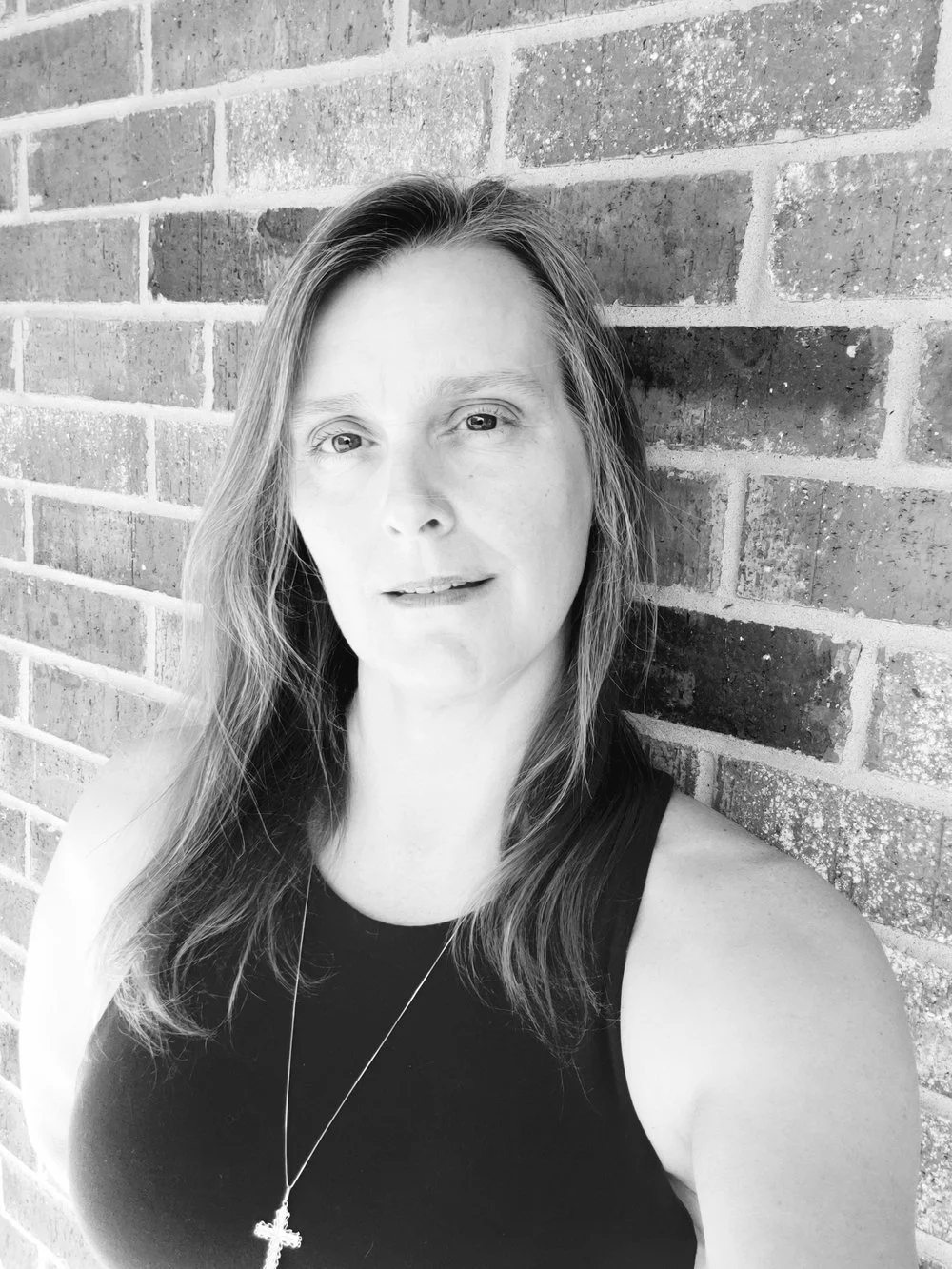 A black and white portrait of a woman with long hair standing against a brick wall, wearing a sleeveless top and a necklace with a cross pendant.