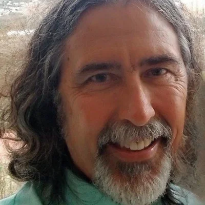 Close-up of a middle-aged man with long, wavy gray hair and a beard, smiling outdoors.