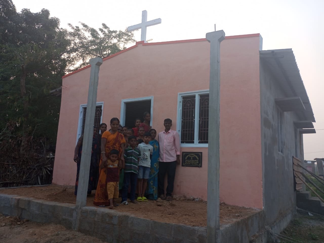 A group of people standing in front of a newly built church with pink walls, blue windows, and a cross on top, outdoors during the daytime.