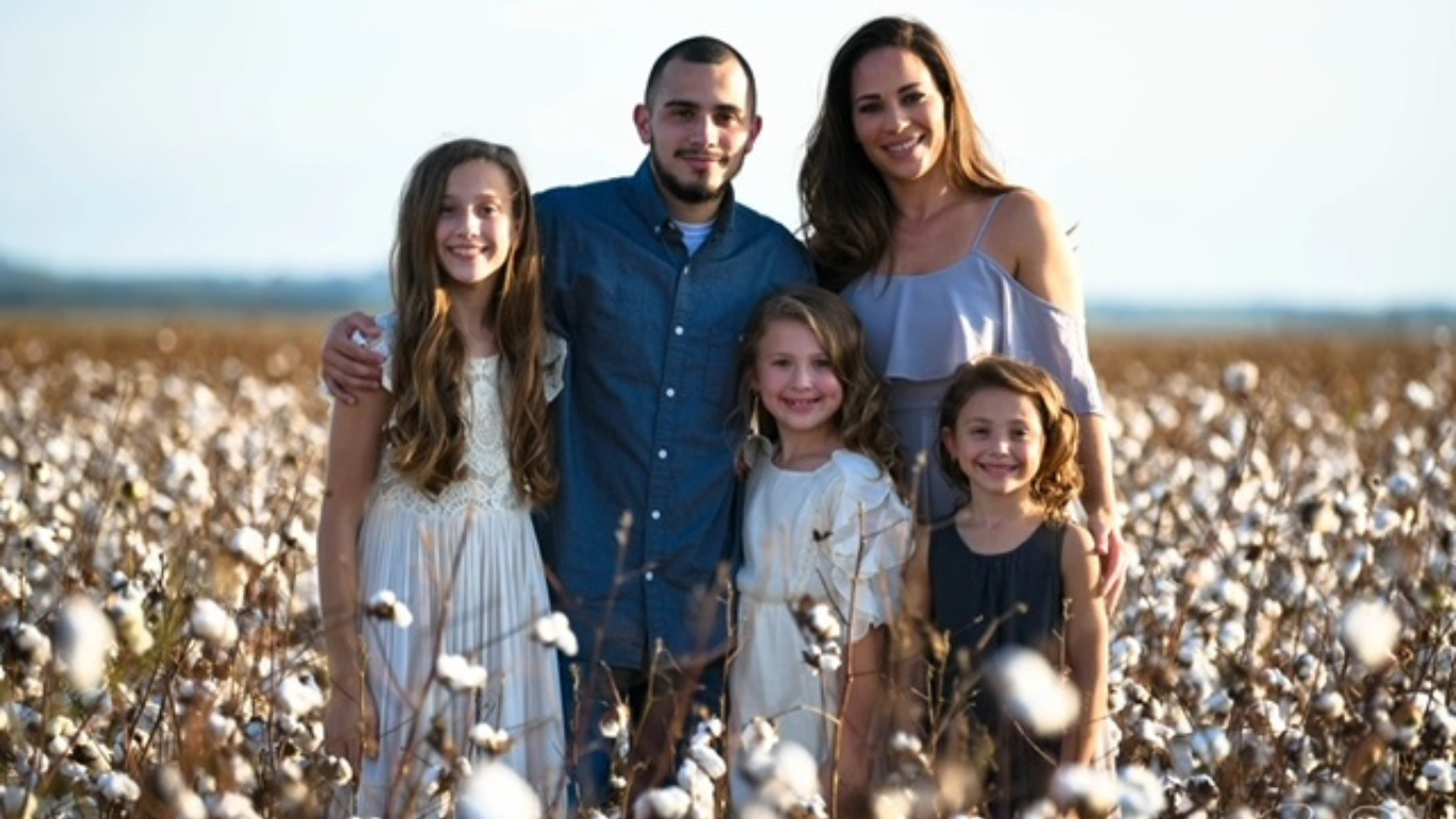 A family of five standing in a cotton field during daytime, smiling at the camera. The group includes a young girl in a white dress, a man in a blue shirt, a woman in a light purple off-shoulder top, and two younger girls in dark and white dresses.