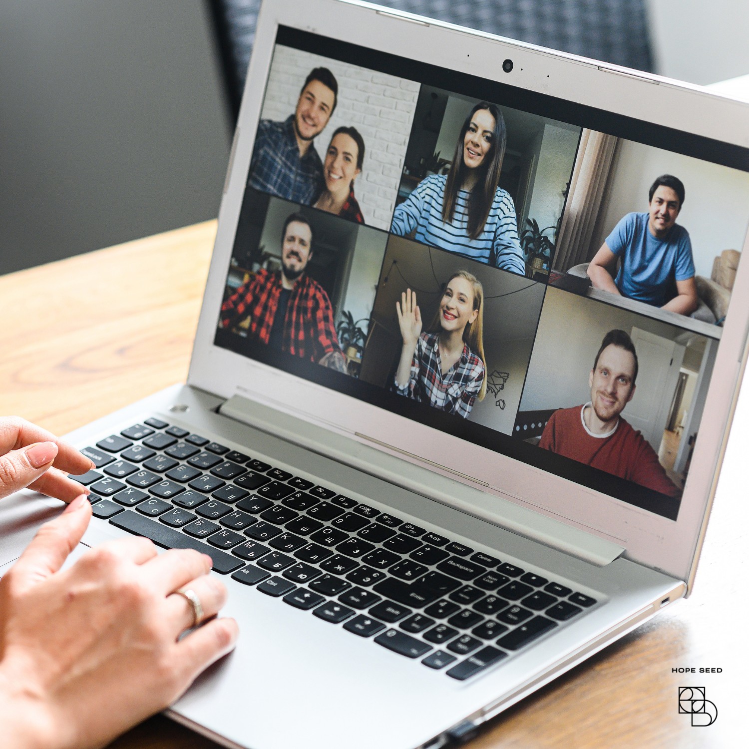 A group of six people are having a video call on a laptop, appearing happy and waving.