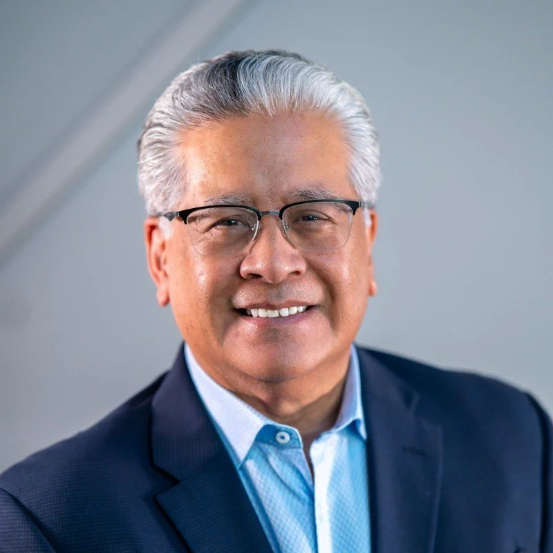 Close-up portrait of a middle-aged man with gray hair and glasses, smiling, wearing a dark blazer and light blue shirt, against a plain background.