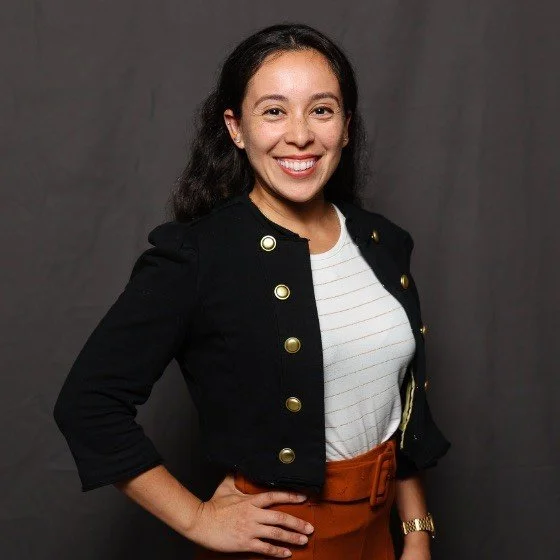 A young woman smiling and posing with her hand on her hip in front of a gray background. She has dark, wavy hair and is wearing a black jacket with gold buttons, a white striped shirt, and brown pants.