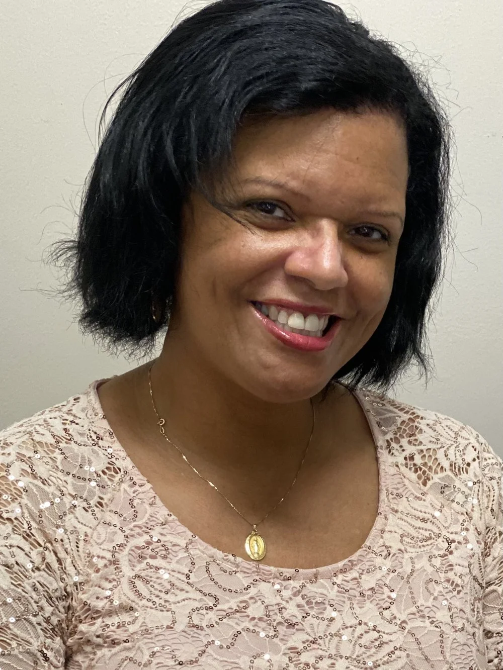 A woman with black hair smiling, wearing a pink lace top and a gold necklace with a pendant, standing against a plain light-colored background.