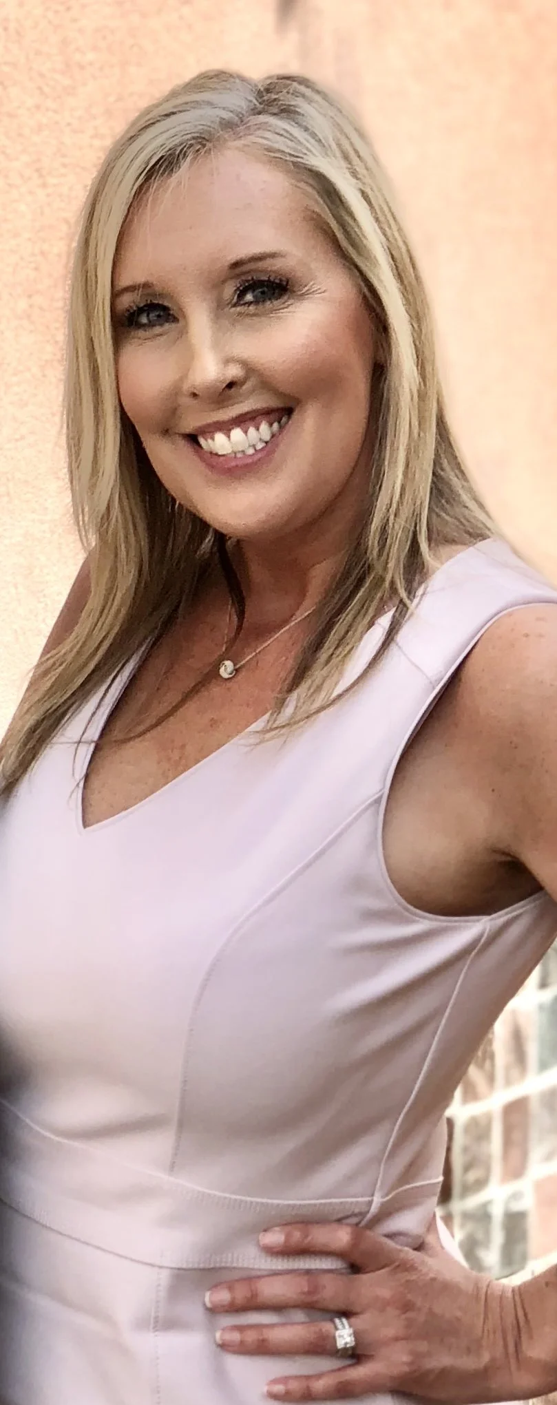 A smiling woman with blonde hair and blue eyes wearing a sleeveless light pink dress, a necklace with a small pendant, and a ring on her left hand, standing with her hand on her hip in front of a neutral background.