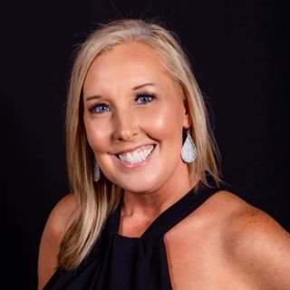 Smiling woman with blonde hair wearing statement earrings and a black top against a dark background.