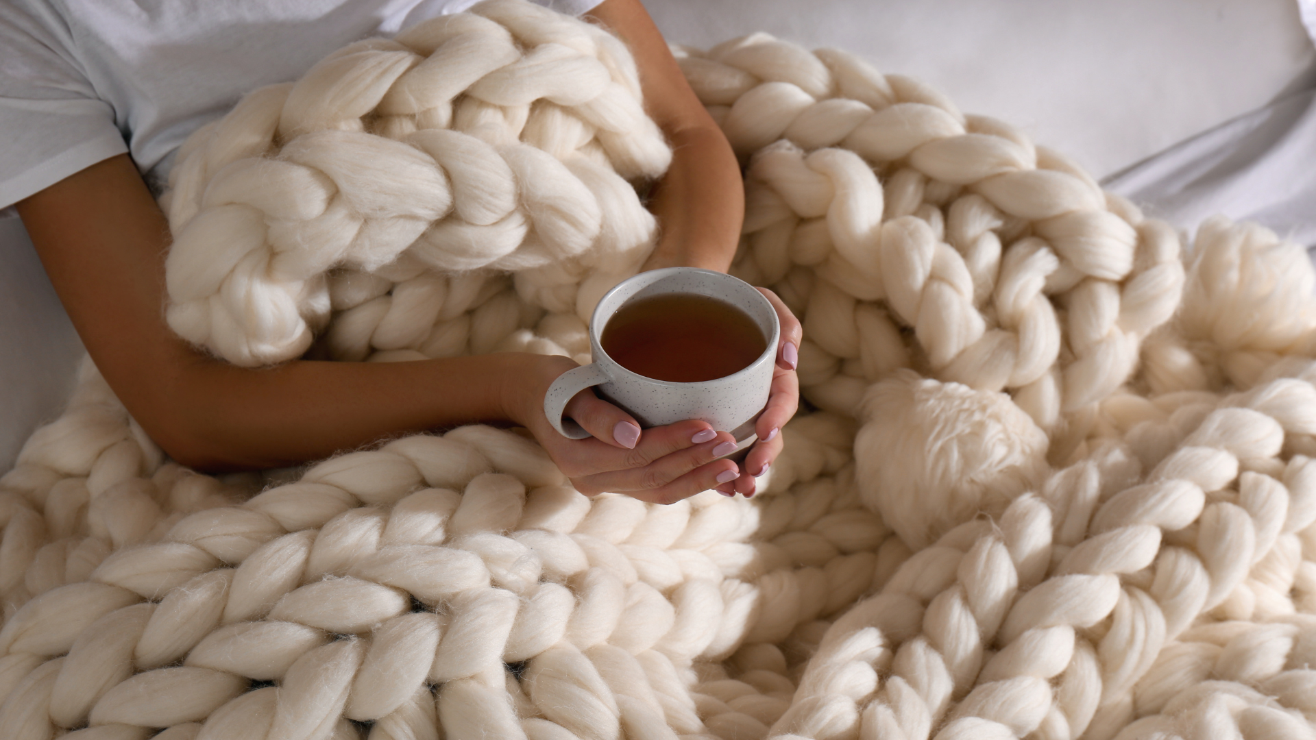 Person holding a white ceramic mug filled with tea, resting on a large, chunky cream-colored knit blanket.