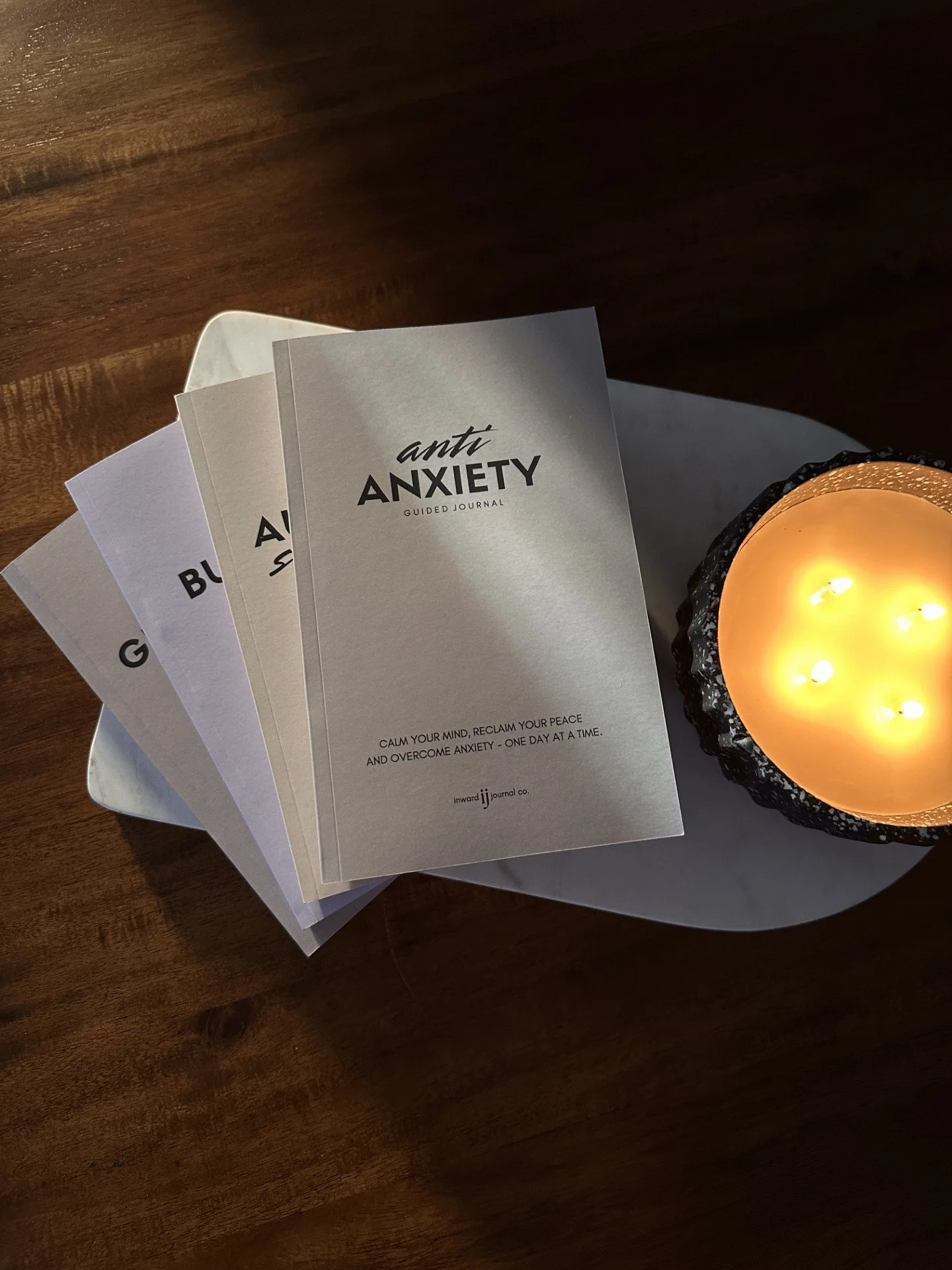 guided journals stacked neatly in a fan pattern, with anti anxiety guided journal on top, sitting on a marble board on a wood table with a lit candle beside them