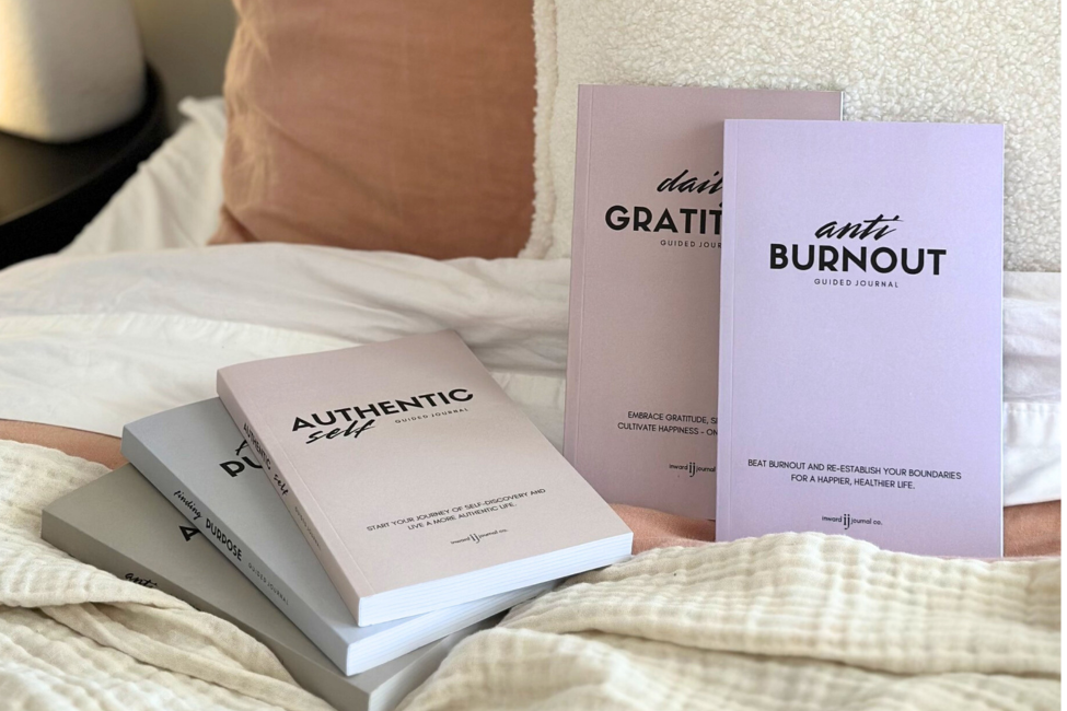 A bed with white and beige bedding and pillows, with four guided journal books titled 'Authentic Self,' 'Grat 1,' 'Anti Burnout,' and 'Purpose' standing upright near the head of the bed.